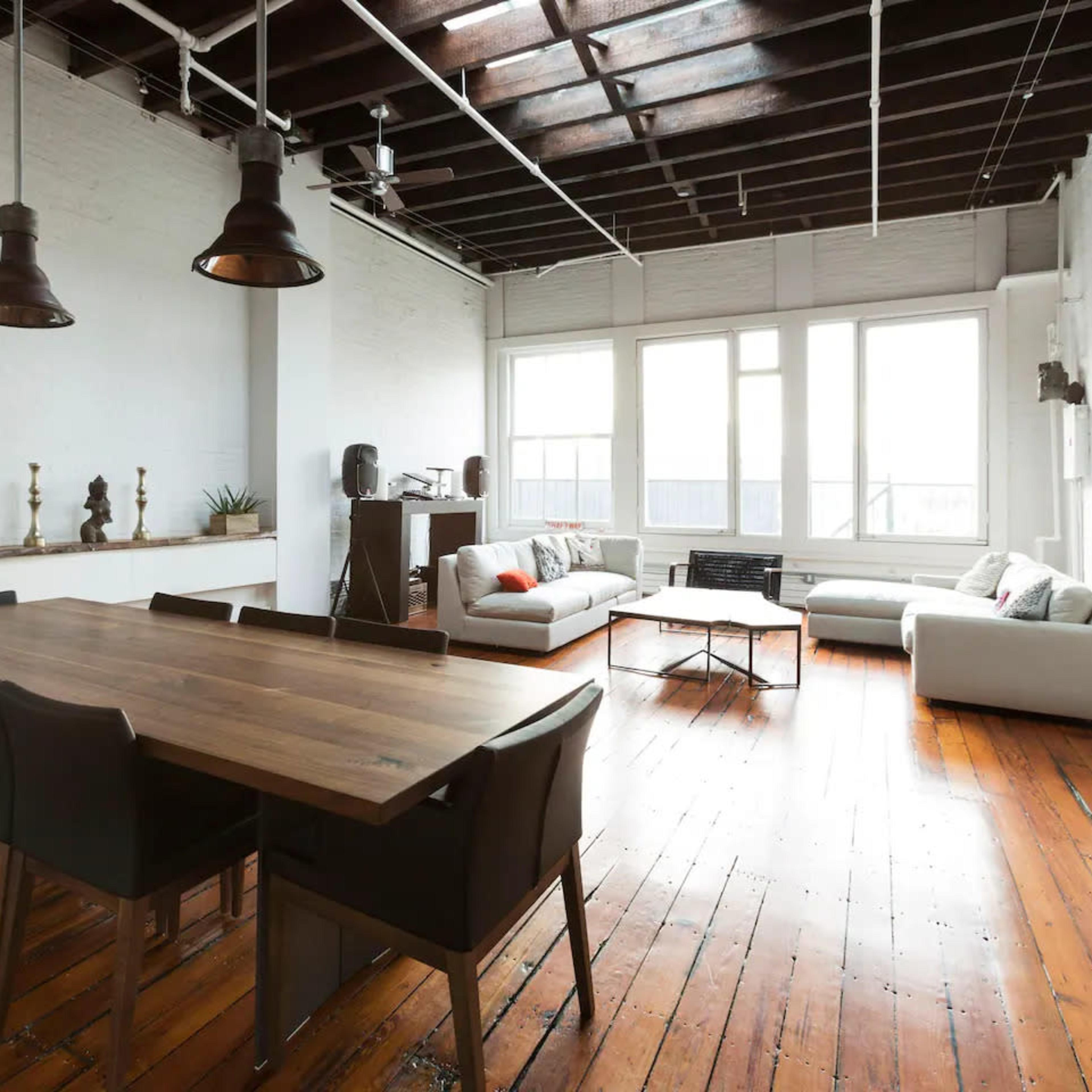 A spacious, well-lit interior featuring a dining table with chairs, a seating area with sofas, and large windows allowing natural light to enter.