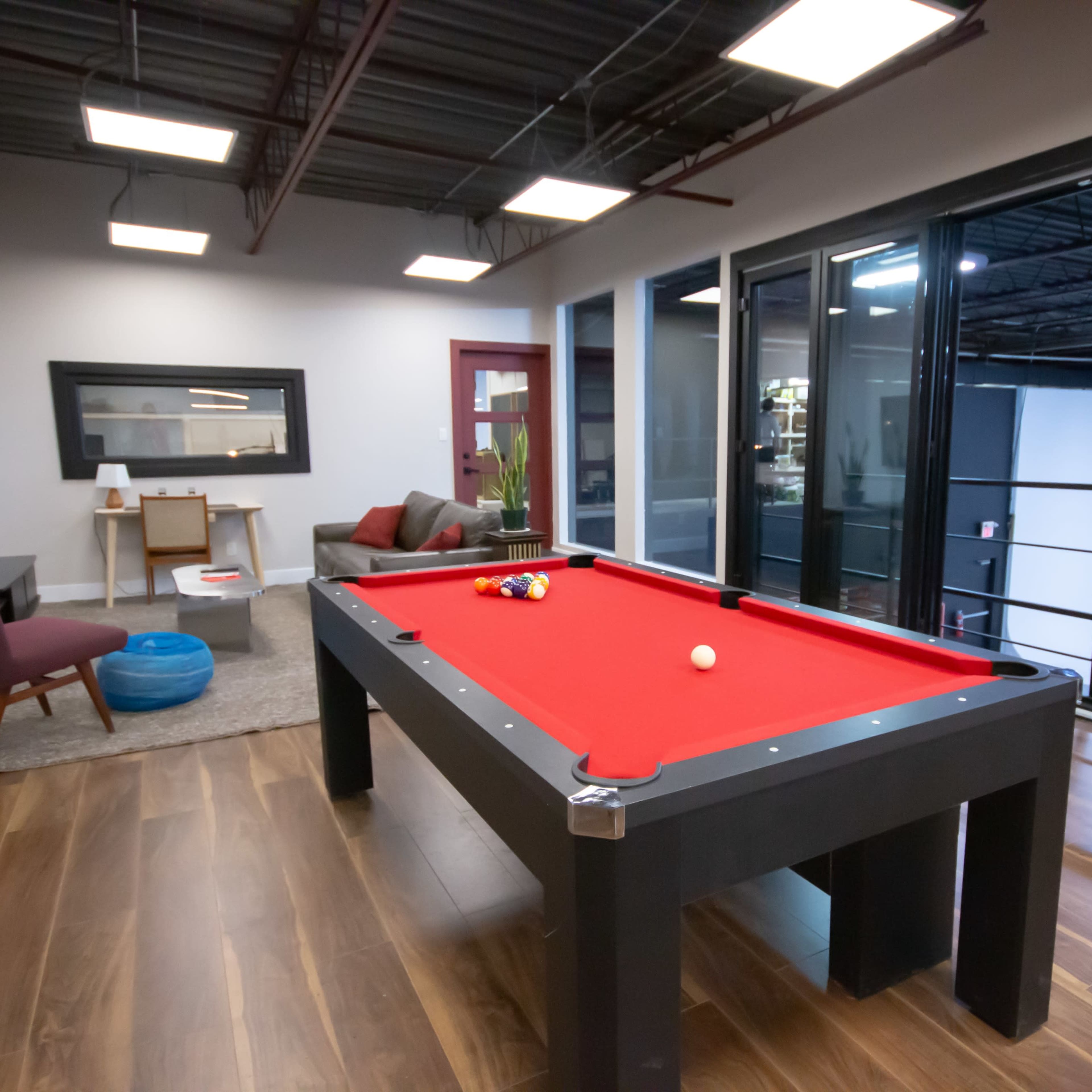 A pool table with a red felt surface is positioned in a room with modern furnishings and large windows.