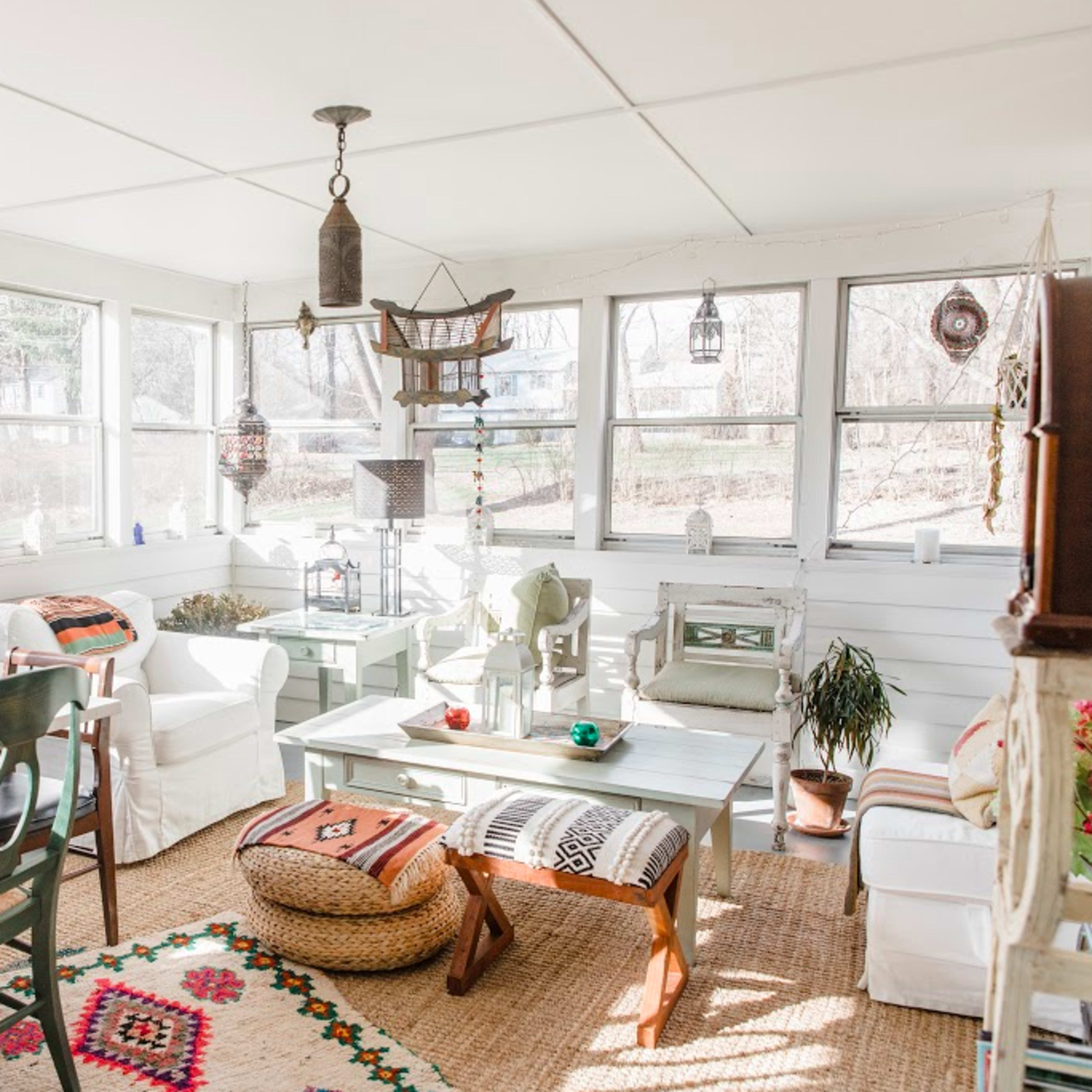 A bright sunroom with white walls, a mix of wooden furniture, and a woven rug, featuring a dining table and various decorative elements.