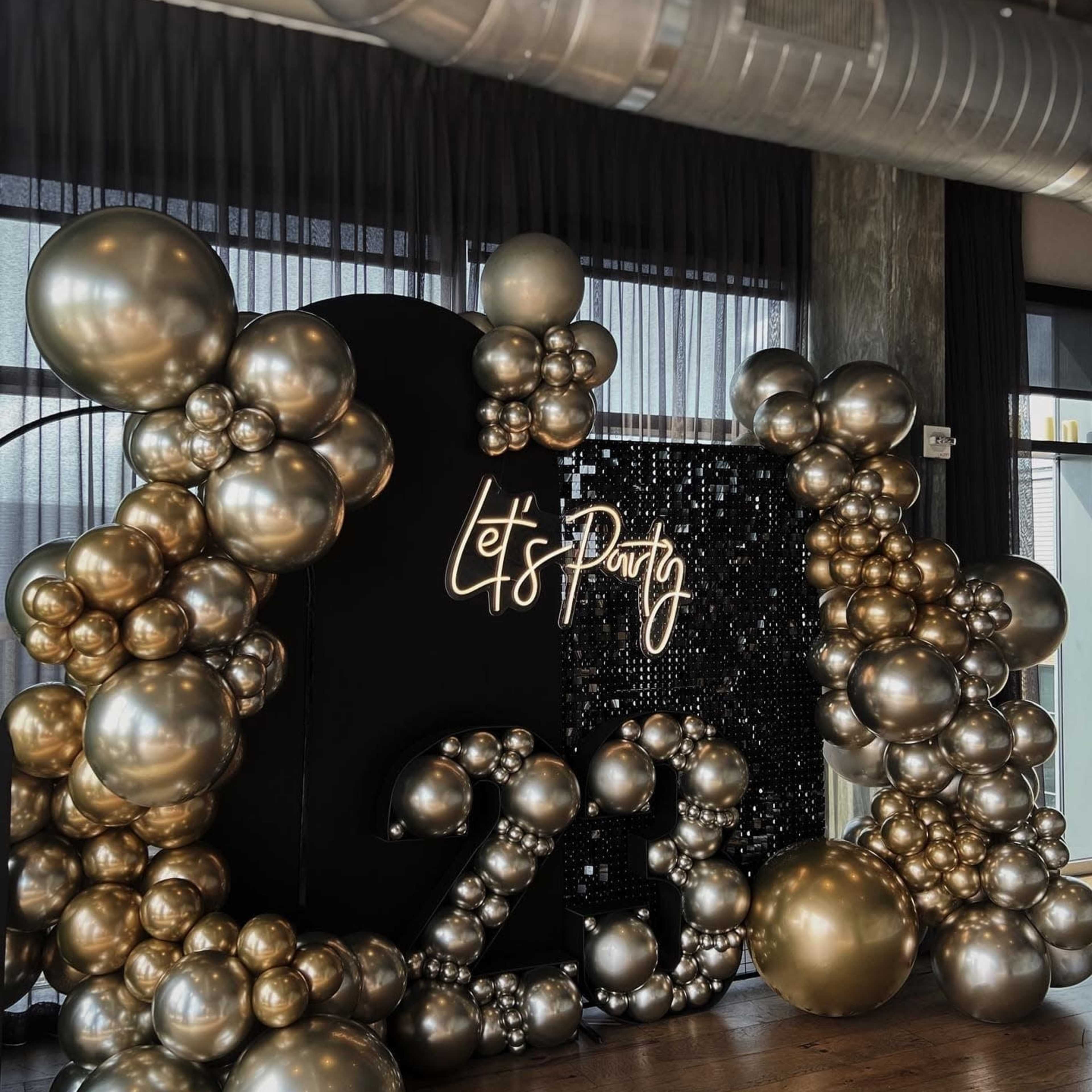 A festive backdrop with large golden and silver balloons arranged around a lit sign that says "Let's Party" and the number "23."