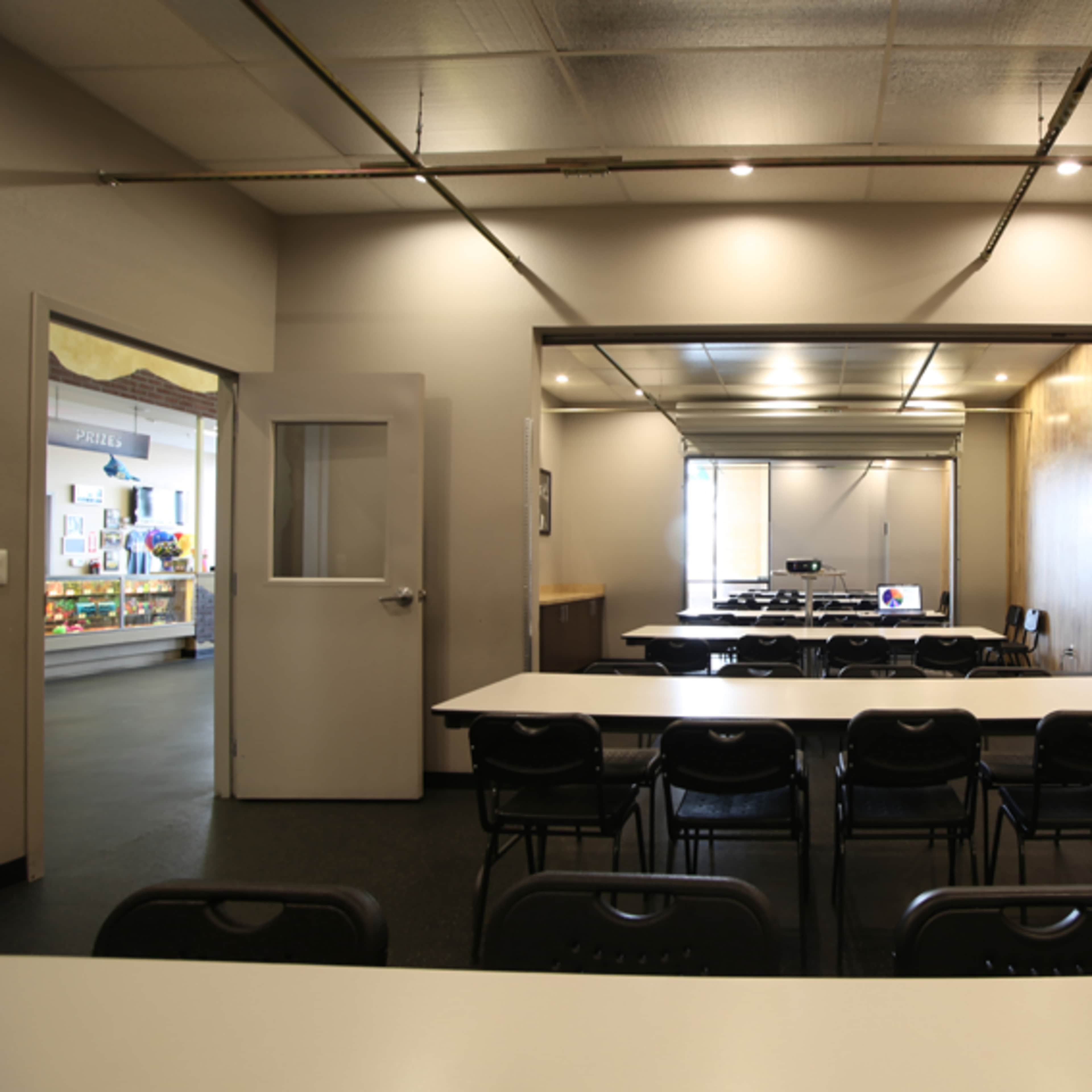 A spacious meeting room featuring several black chairs arranged around white tables, with a large projection screen visible in the background.