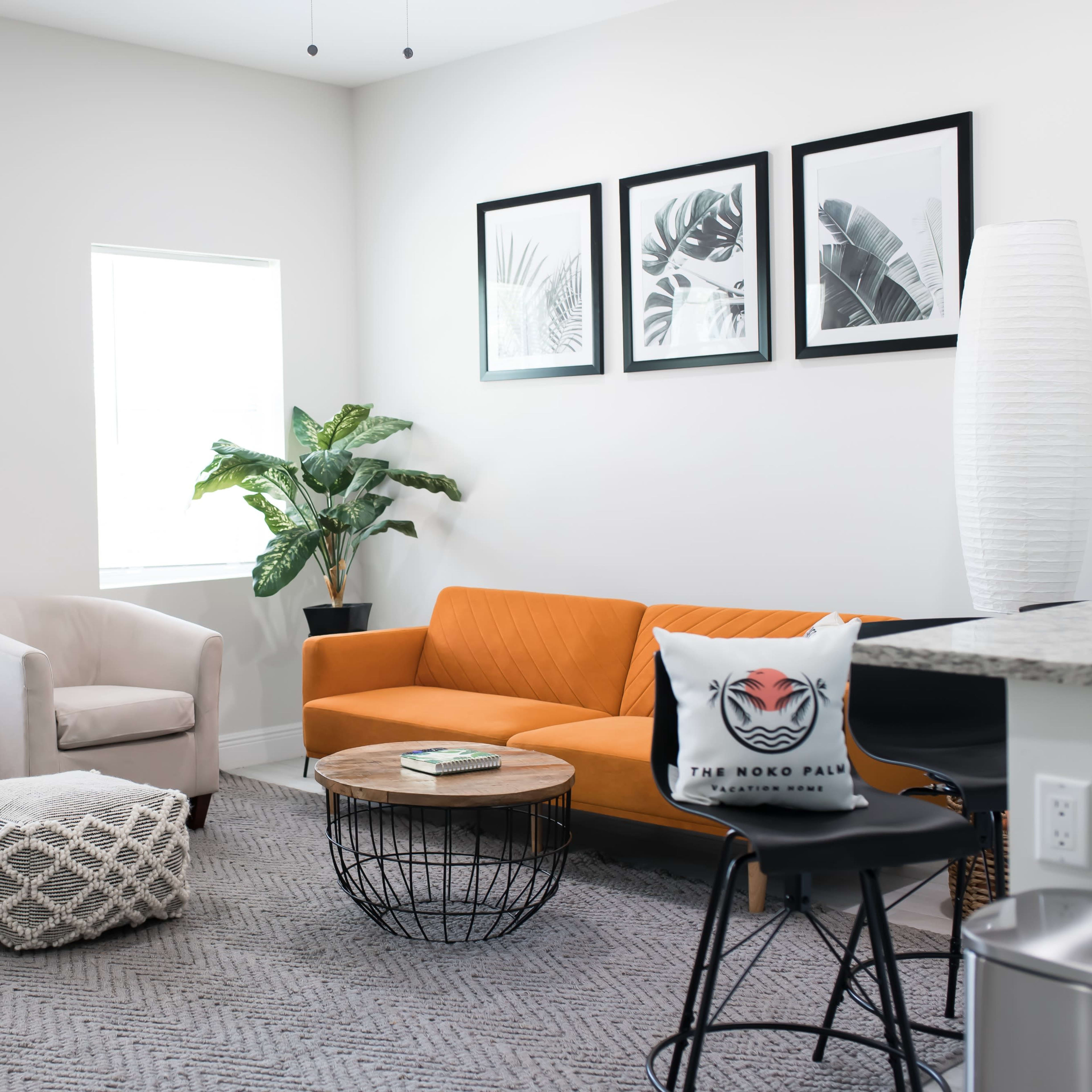 A modern living room with an orange sofa, two light-colored chairs, a wooden coffee table, a patterned rug, and potted plants, illuminated by natural light through two windows.