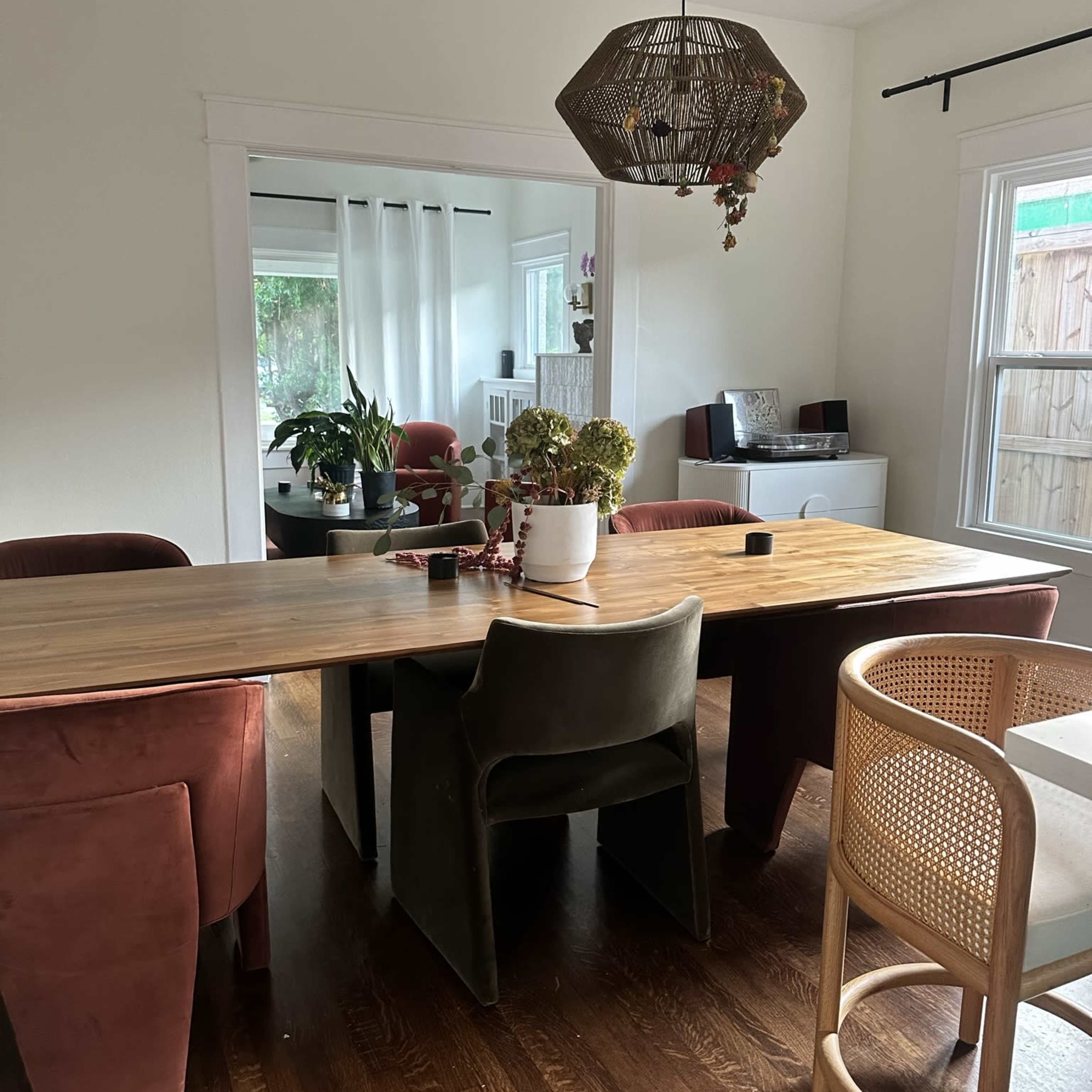 A dining area with a wooden table surrounded by four upholstered chairs and a rattan stool, with a hanging light fixture above and plants visible in the background.