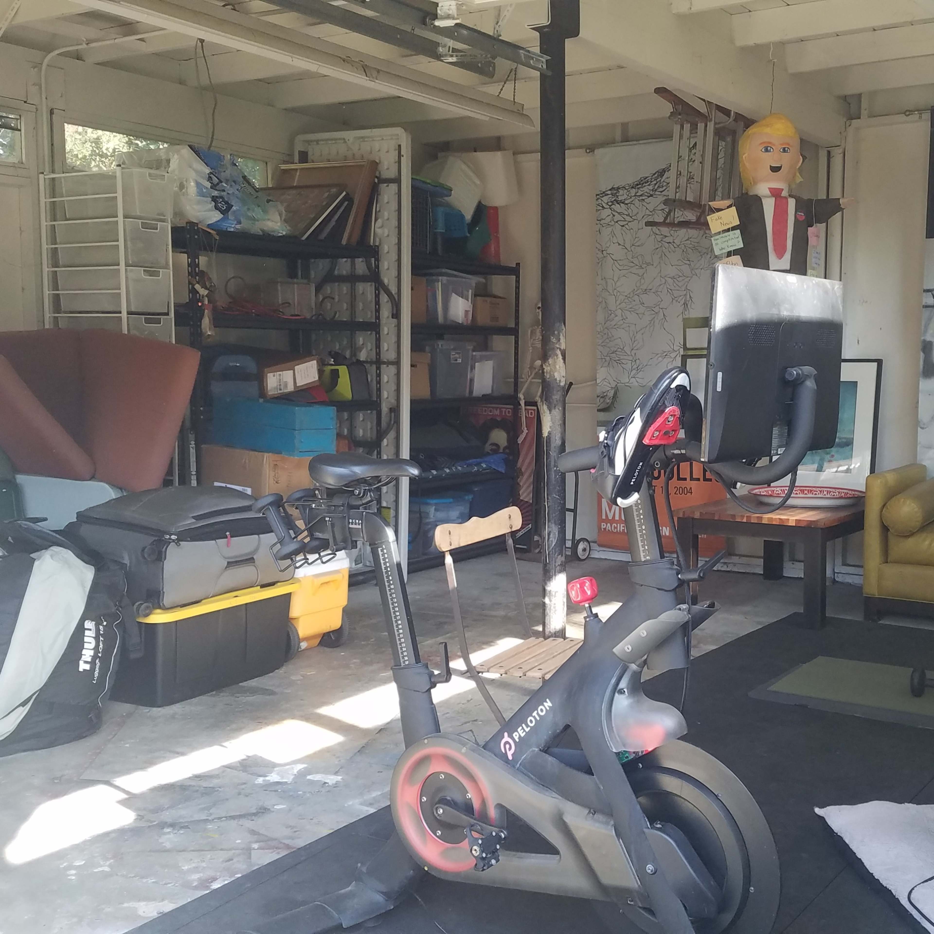 A cluttered garage with exercise equipment, storage boxes, and various pieces of furniture, including a yellow couch and an artistic backdrop.