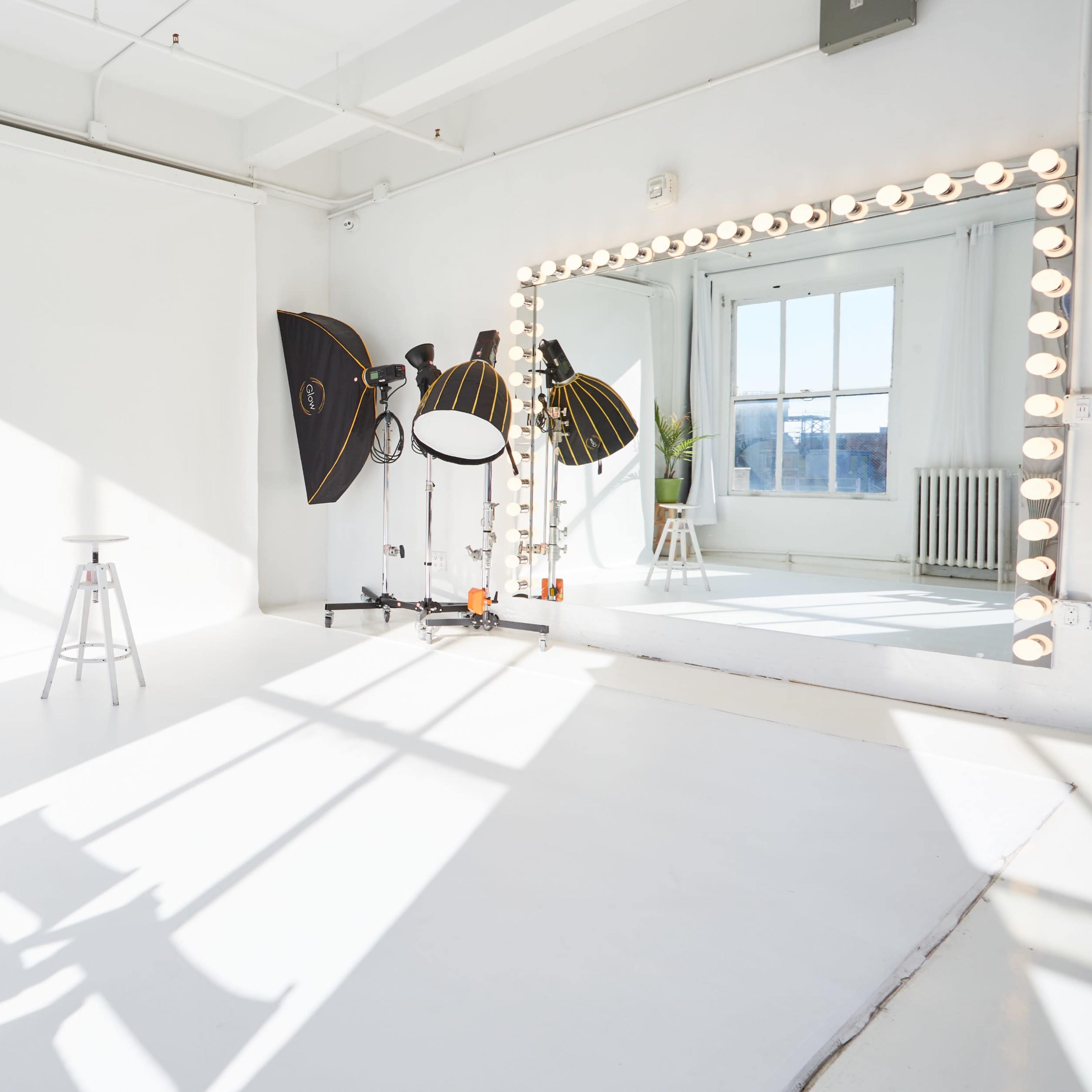 A bright photography studio with a white backdrop, a stool, lighting equipment, and a large mirror framed with bulbs.