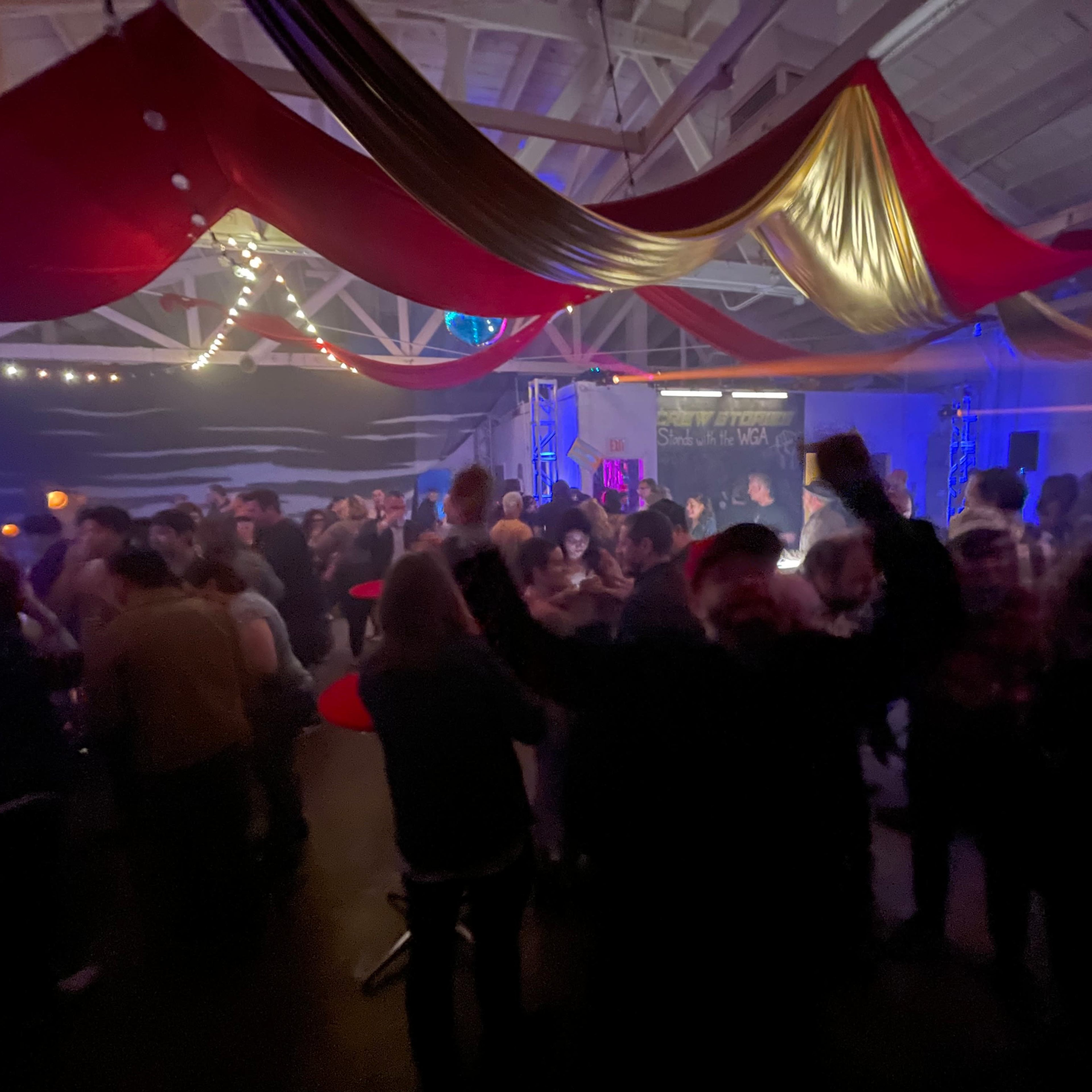A crowded indoor venue features people socializing under draped red and gold fabric with string lights illuminating the scene.