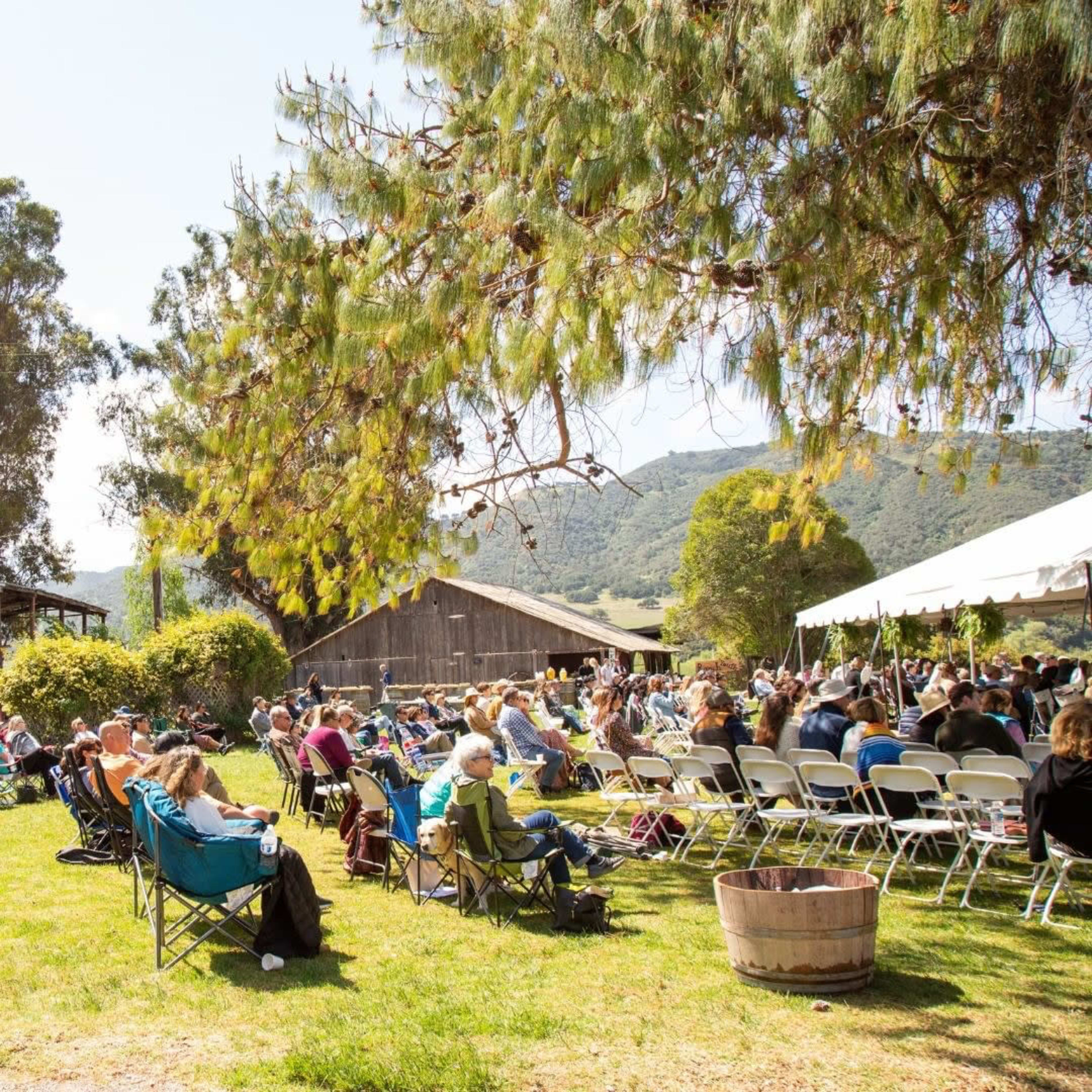 A large group of people sits in folding chairs under a tent on a grassy area, with trees and a barn in the background.