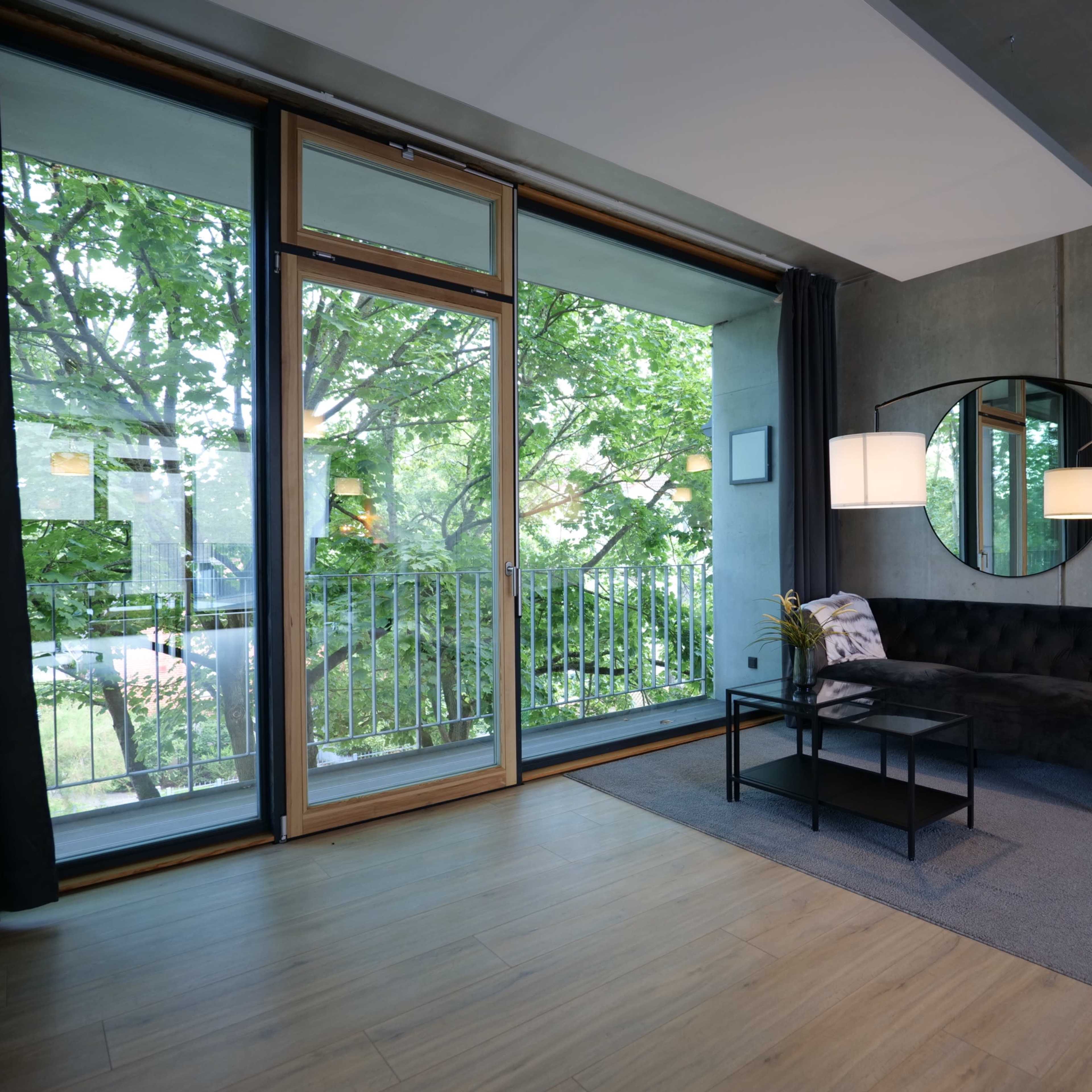 A modern living room featuring a dark sofa, a round mirror, a glass coffee table, and large windows with views of greenery outside.