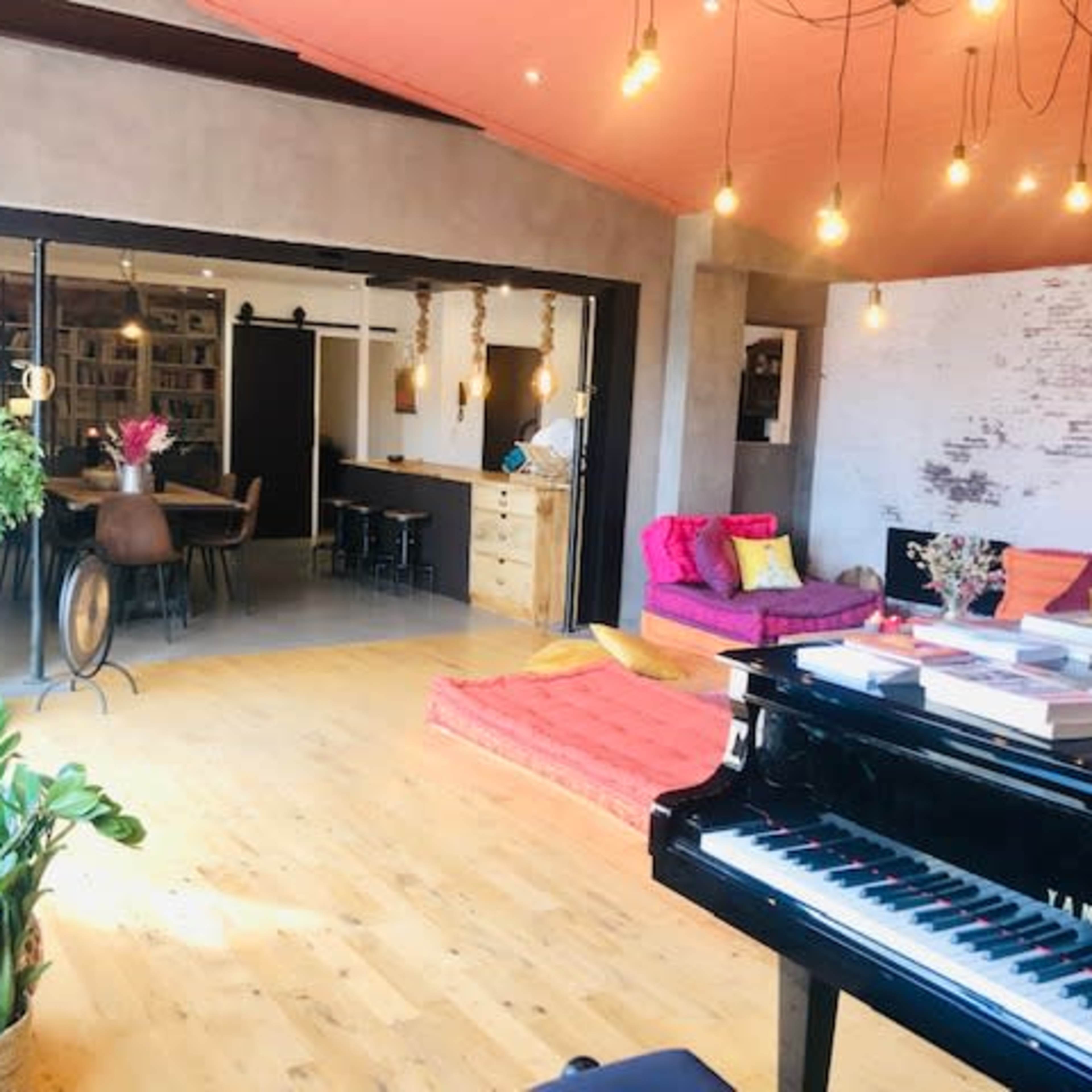 A spacious living area features a grand piano in the foreground, colorful seating arrangements, and an open kitchen in the background.