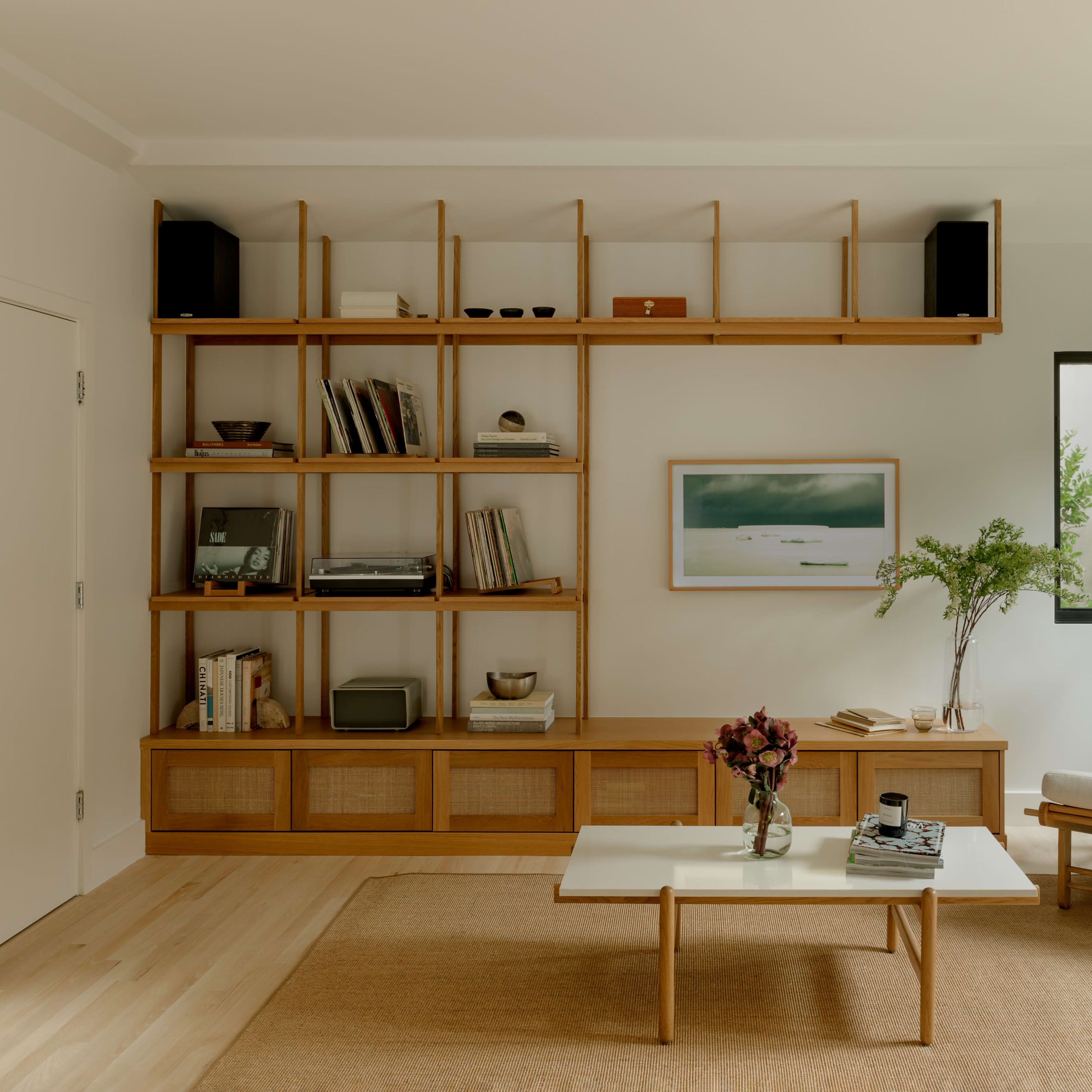 A modern living room with a wooden shelving unit, a white armchair, a coffee table, and large windows allowing natural light to enter.