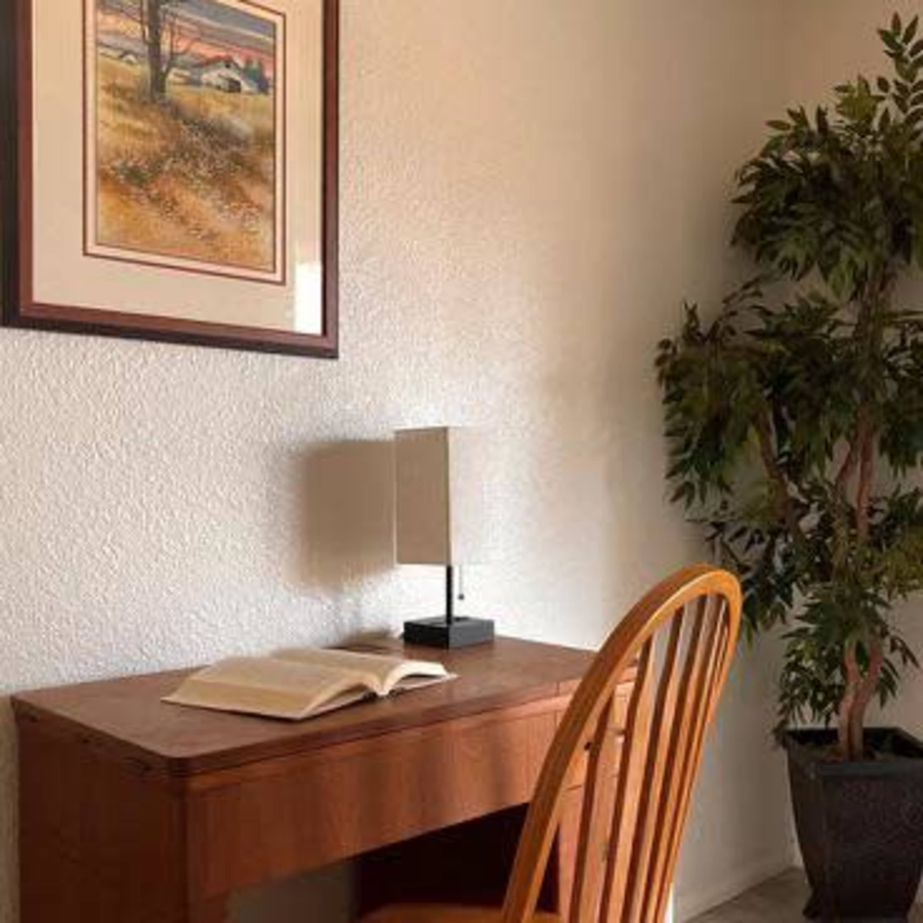 A wooden desk with a chair and an open book sits next to a framed landscape painting and a small lamp, beside a potted plant.