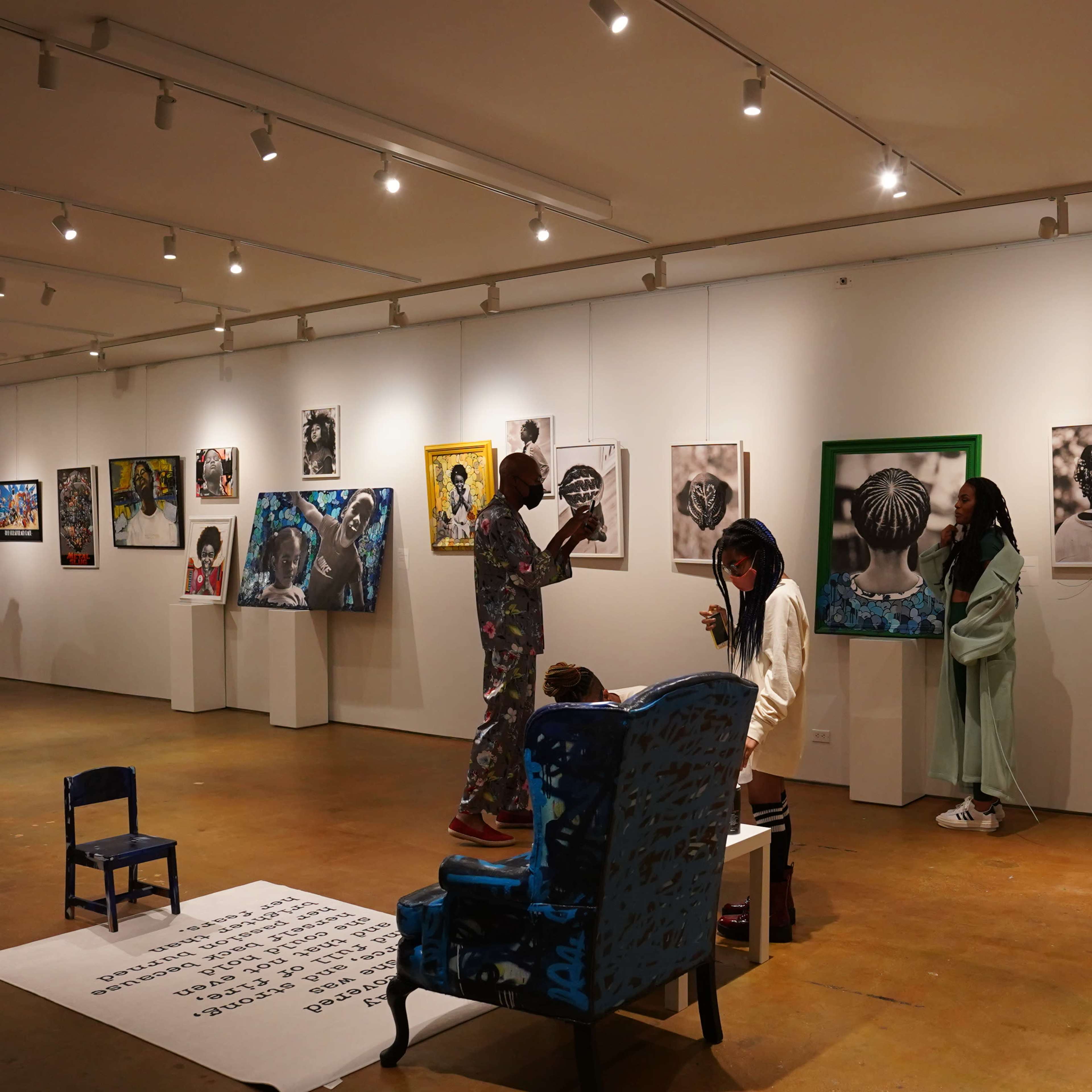 An art gallery is filled with people viewing various framed artworks on the walls, while a small blue chair and a patterned sofa are positioned in the center of the room.