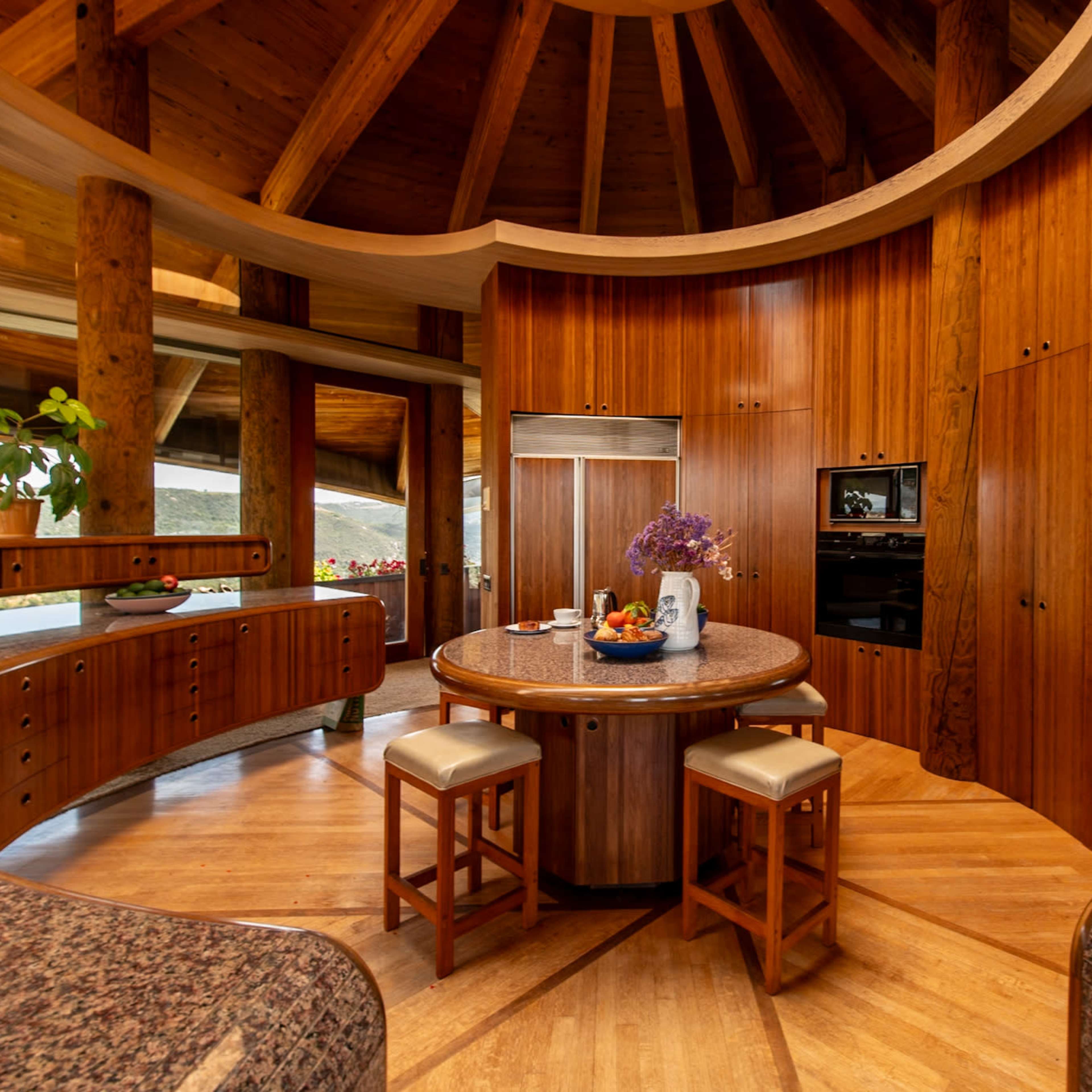 A round kitchen featuring wooden cabinetry, a circular island with stools, and large windows allowing natural light.