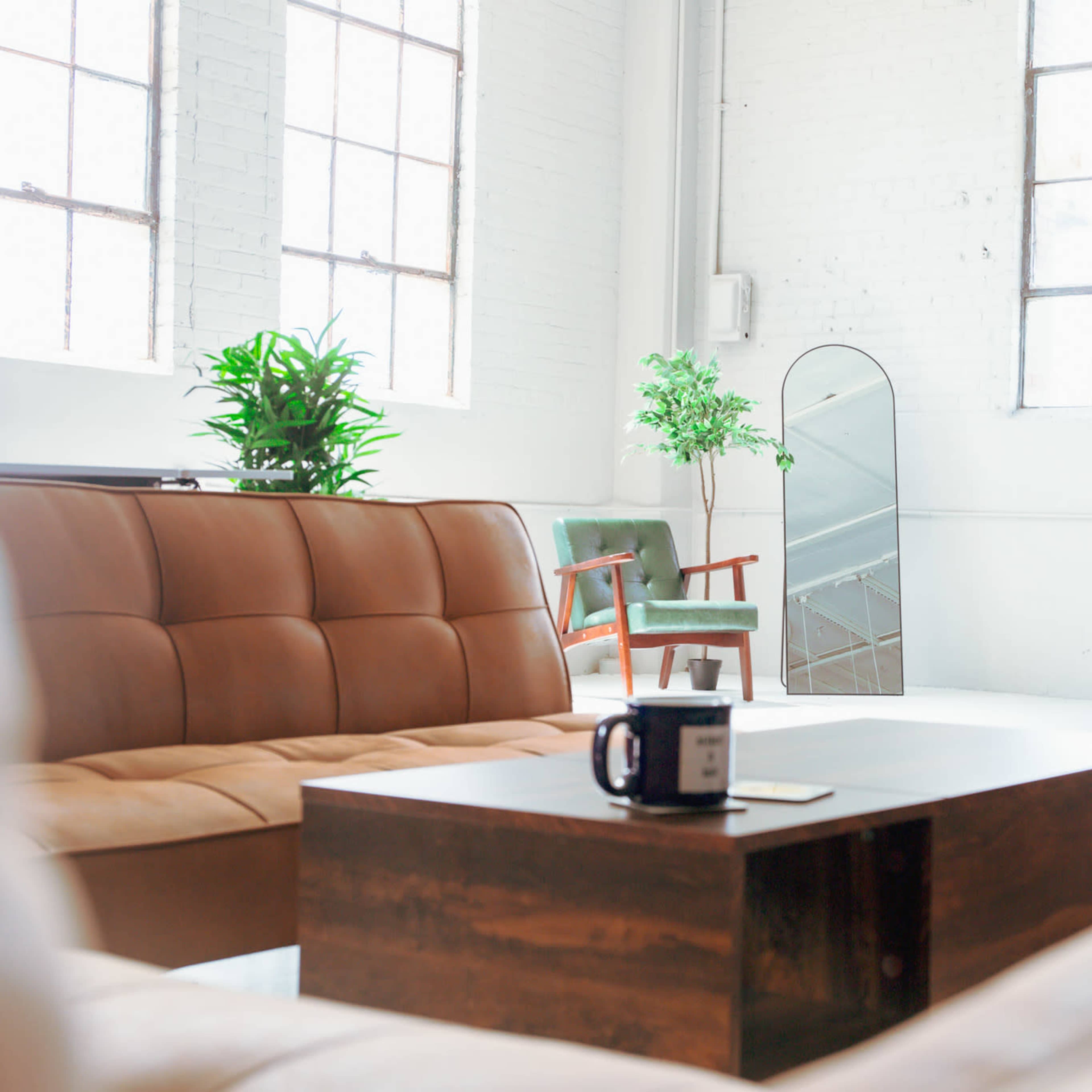A spacious, well-lit living area features brown leather sofas, a wooden coffee table with a mug, and large windows alongside a green plant and an ornate mirror.