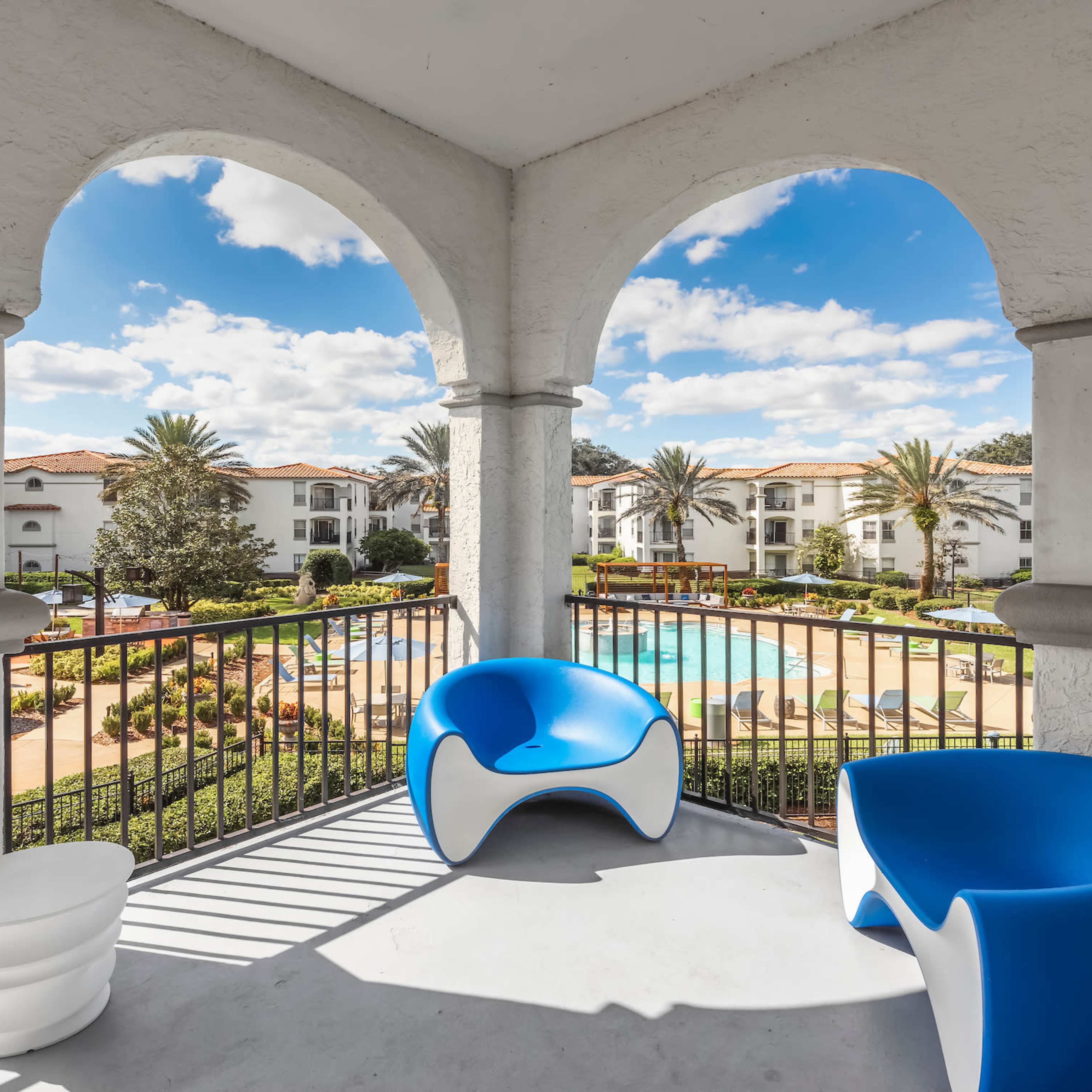 A covered balcony with modern blue and white chairs overlooking a landscaped area with a swimming pool and palm trees in the background.