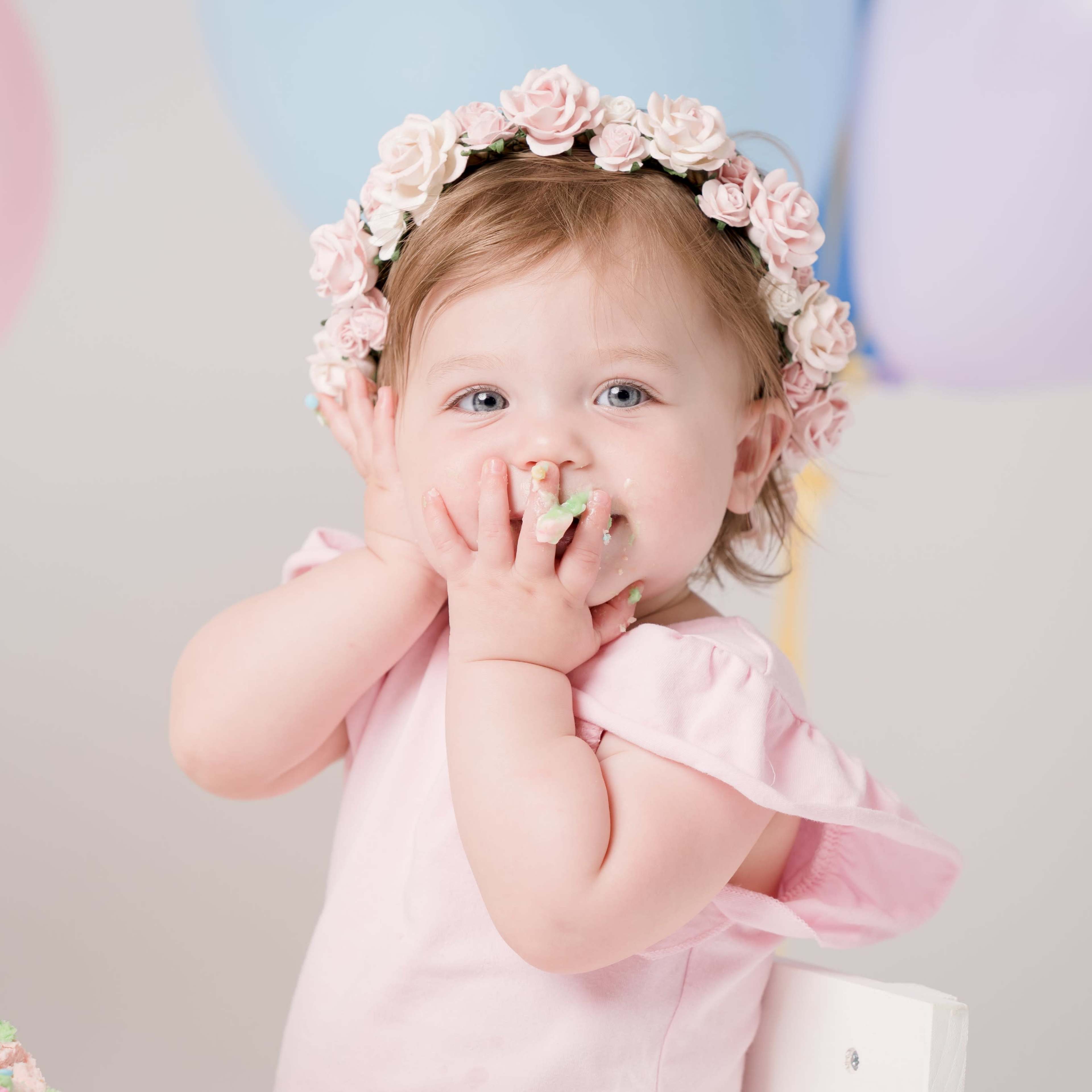 A toddler in a pink dress with a floral crown smiles while eating cake in front of colorful balloons.