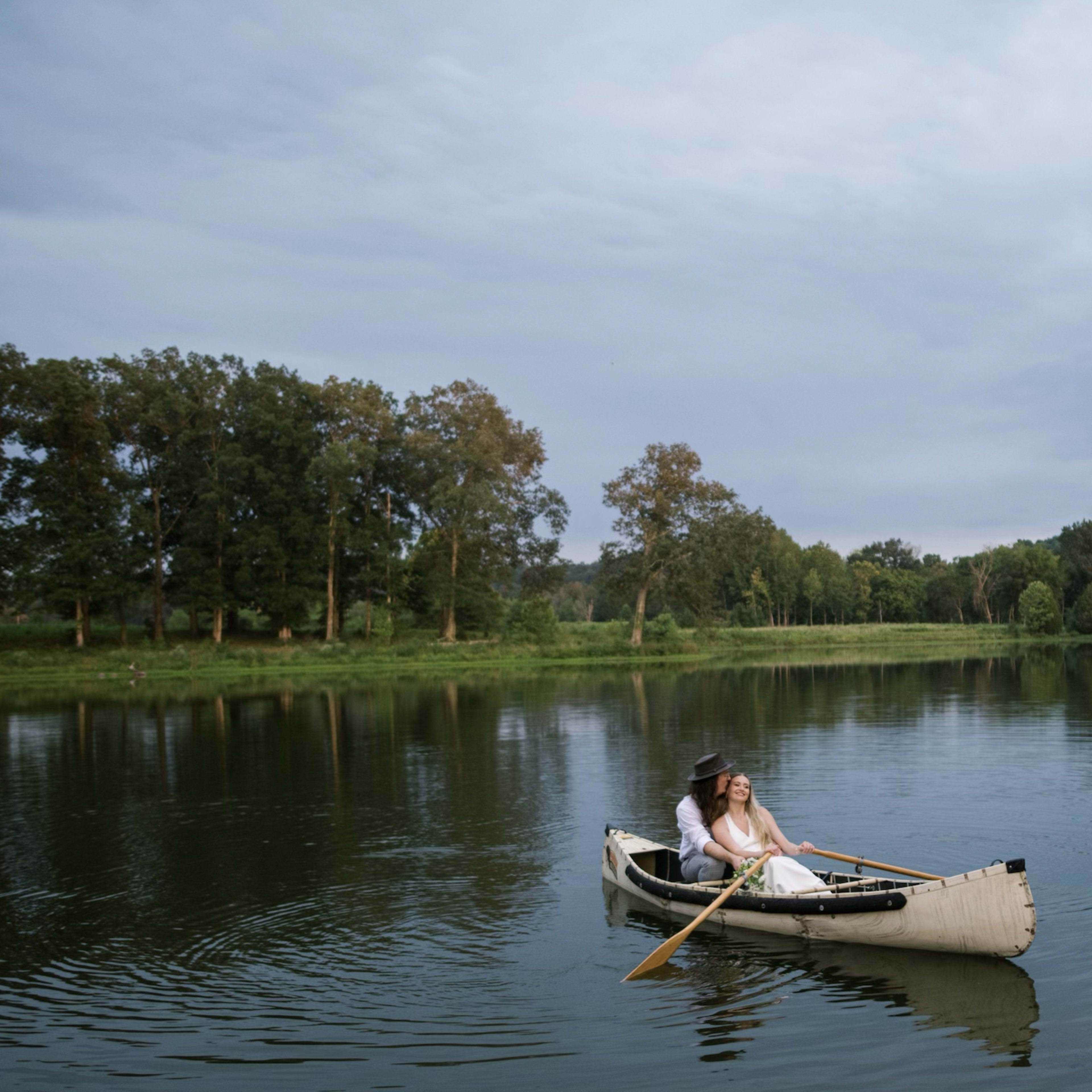 A couple paddles a canoe on a calm lake surrounded by trees and gentle hills.