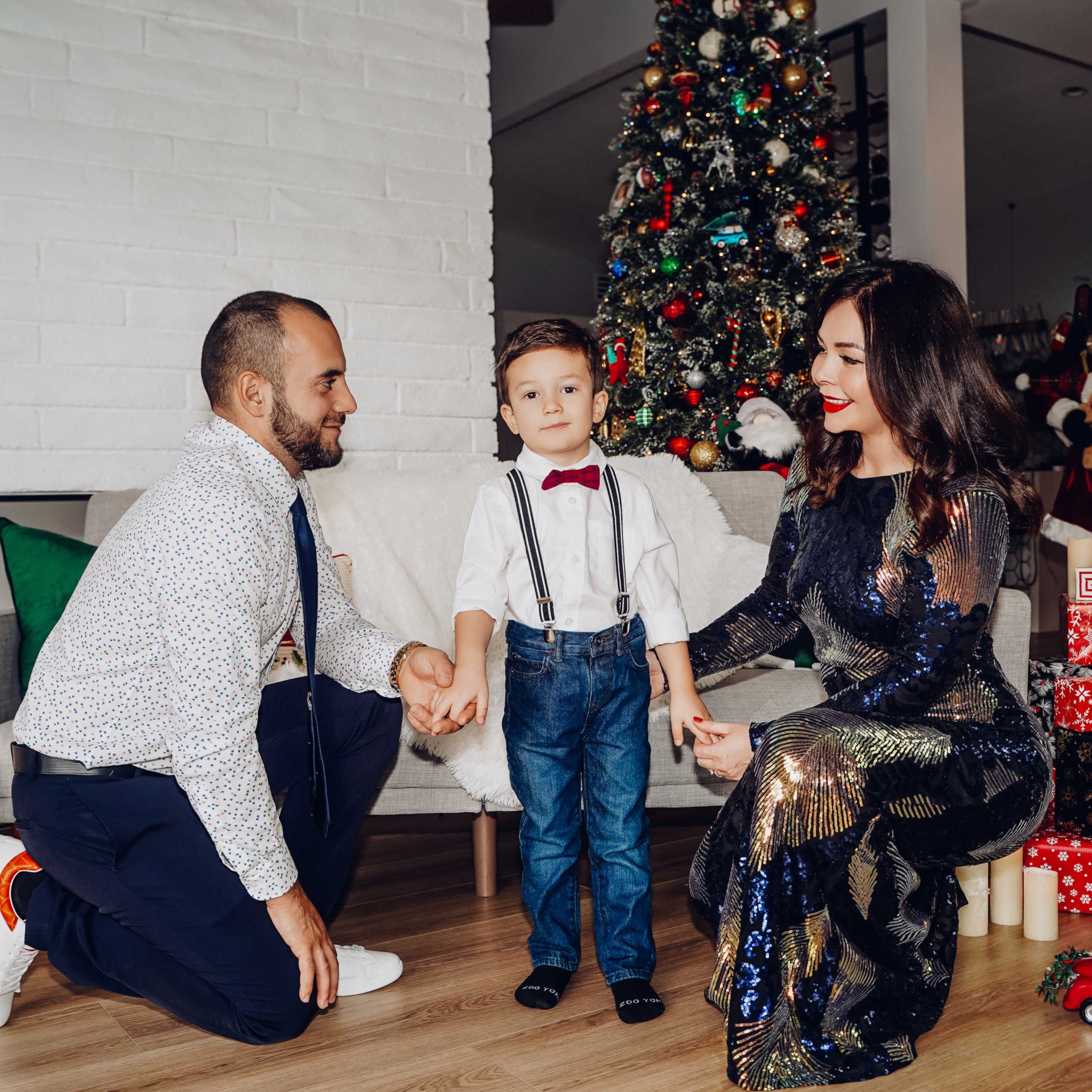A family is gathered in a festively decorated living room with a Christmas tree and presents, holding hands while smiling together.
