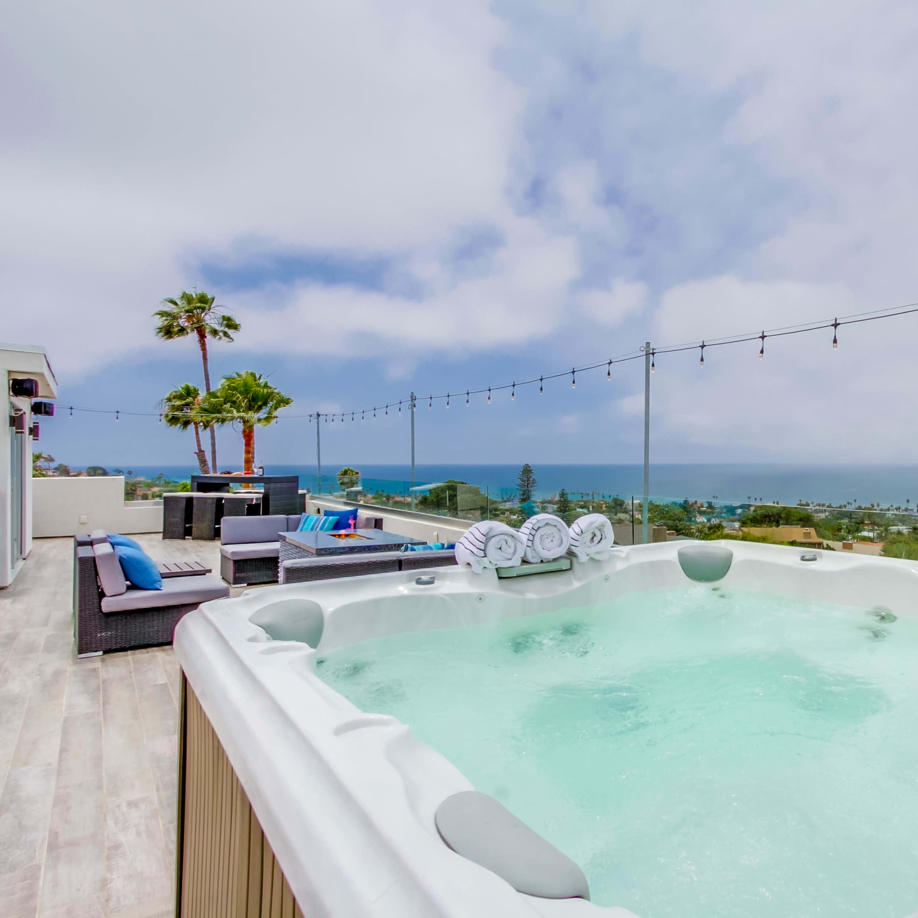 A hot tub is positioned on a deck overlooking a coastal view, with string lights above and seating areas nearby.
