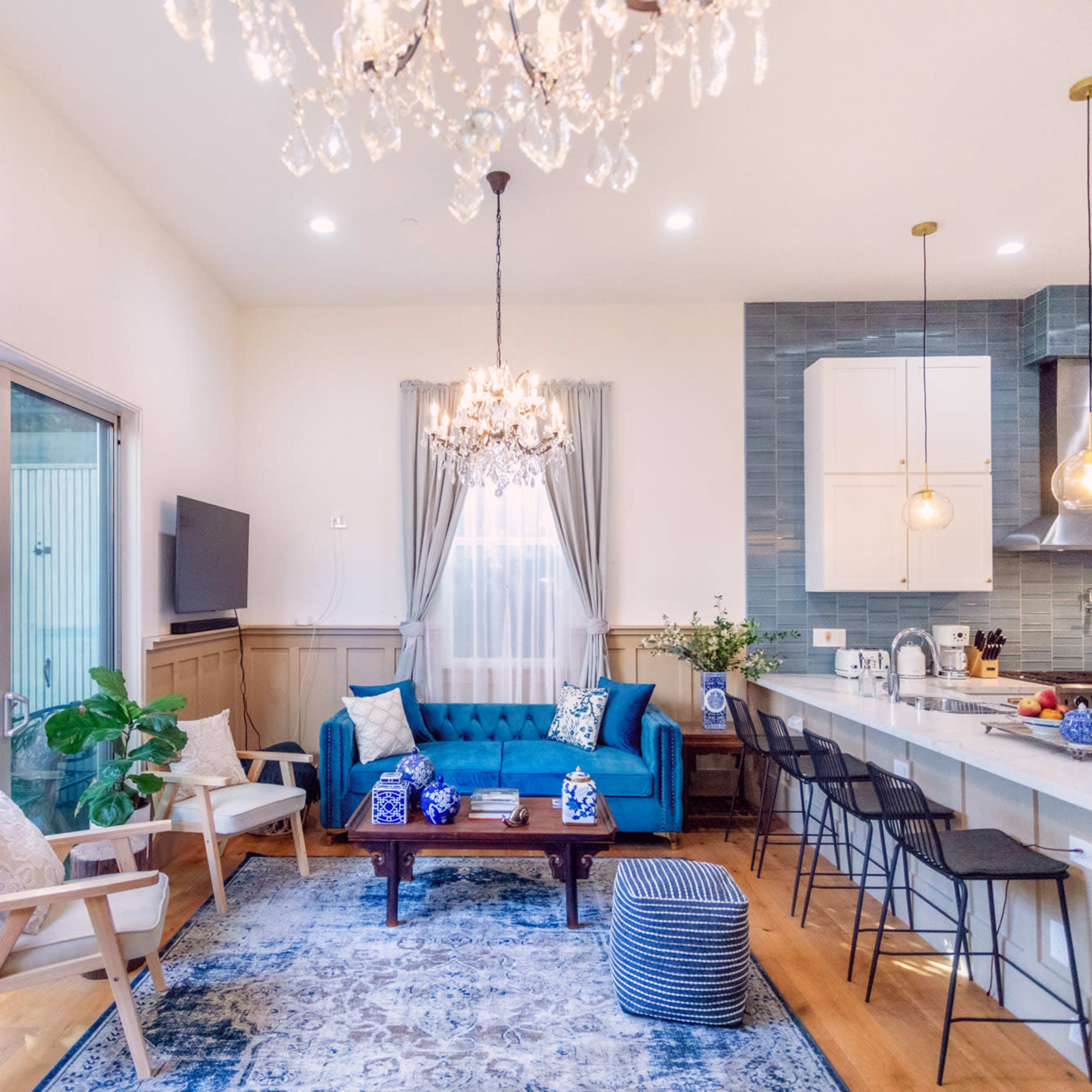 A bright living area features a blue sofa, a coffee table, and a kitchen with modern appliances, all illuminated by a chandelier and pendant lights.