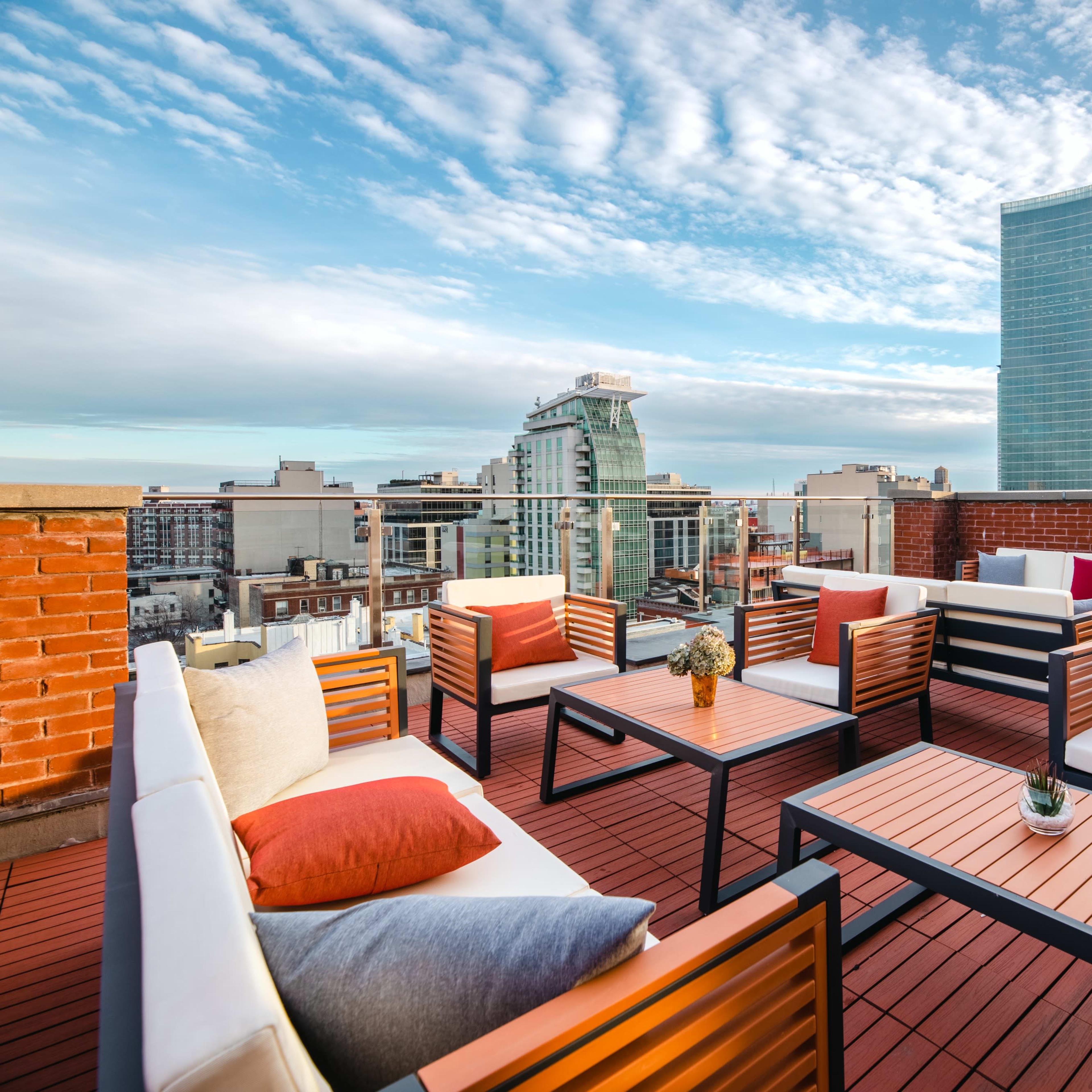 A rooftop terrace featuring modern seating arrangements with tables, overlooking a city skyline under a partly cloudy sky.