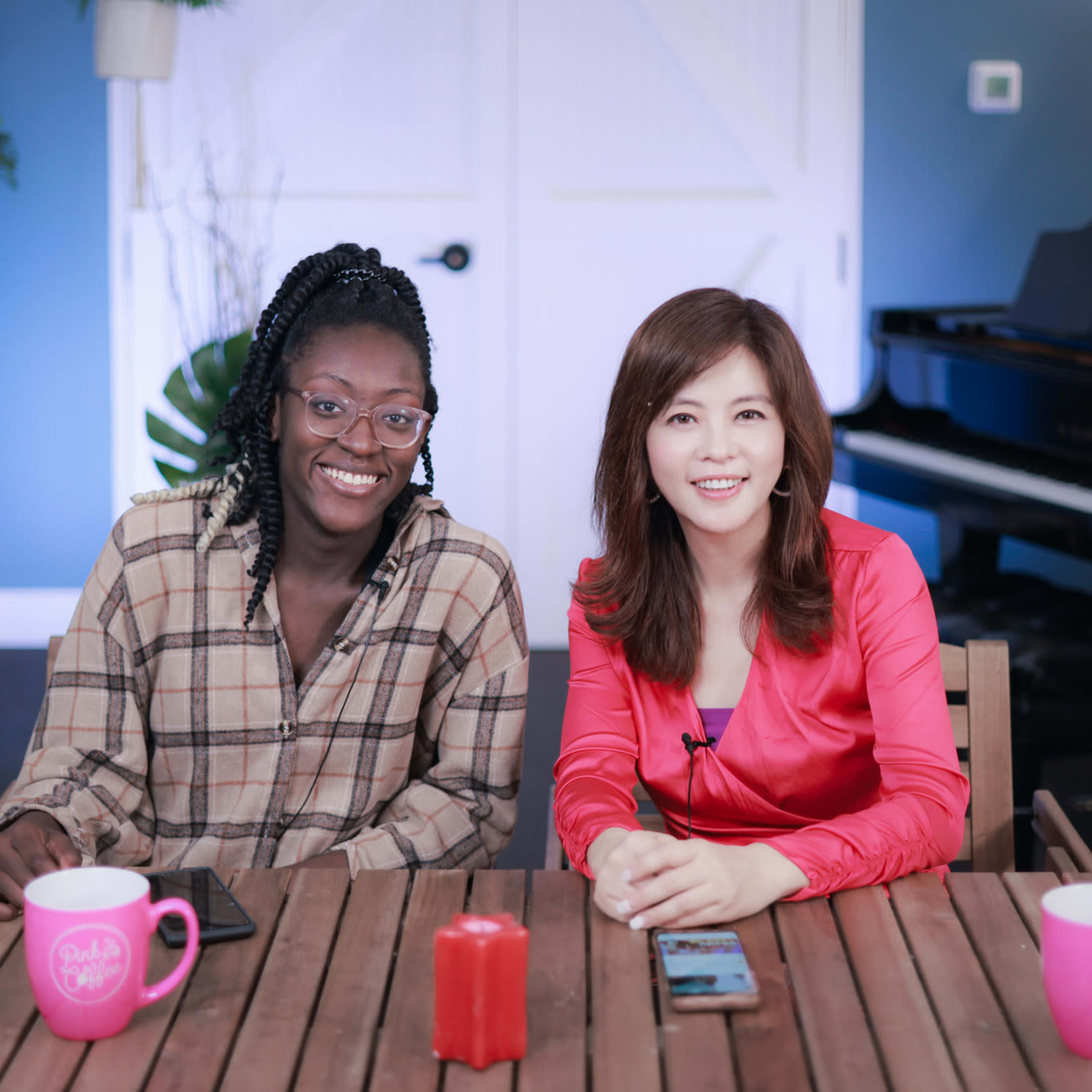 Two women sit at a wooden table, smiling, with coffee mugs in front of them, and a piano in the background.