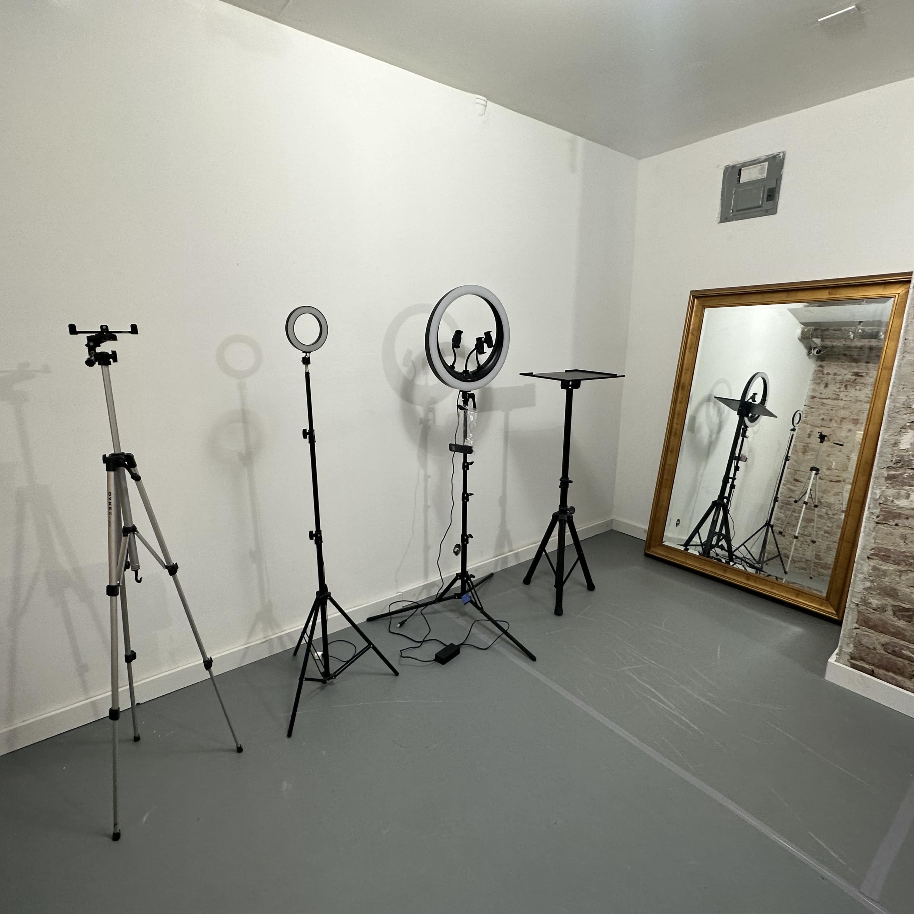 A studio room with a gray floor, featuring tripods, a ring light, and a large mirror against a textured brick wall.