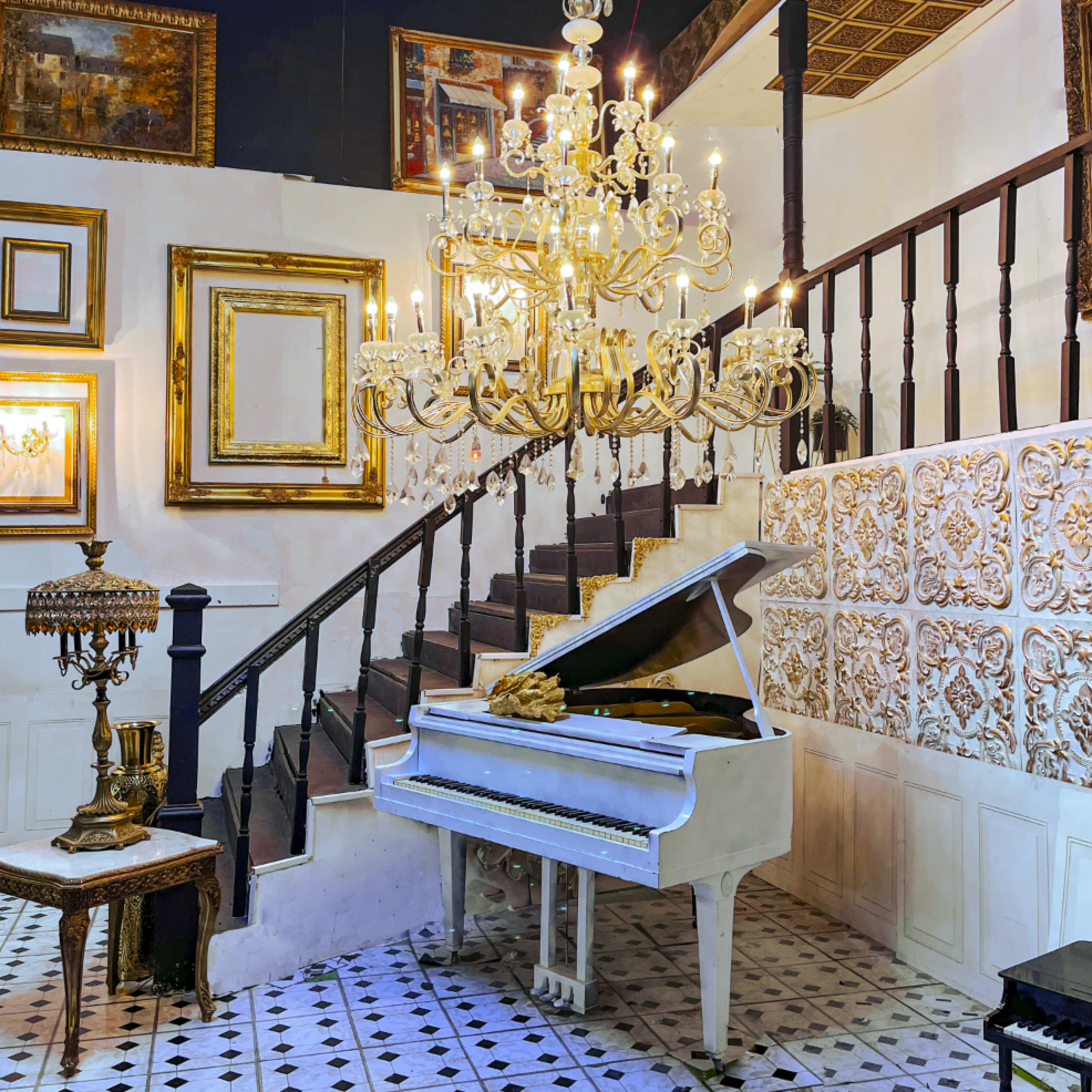 A white grand piano sits near a staircase adorned with a chandelier and framed artworks on the walls.