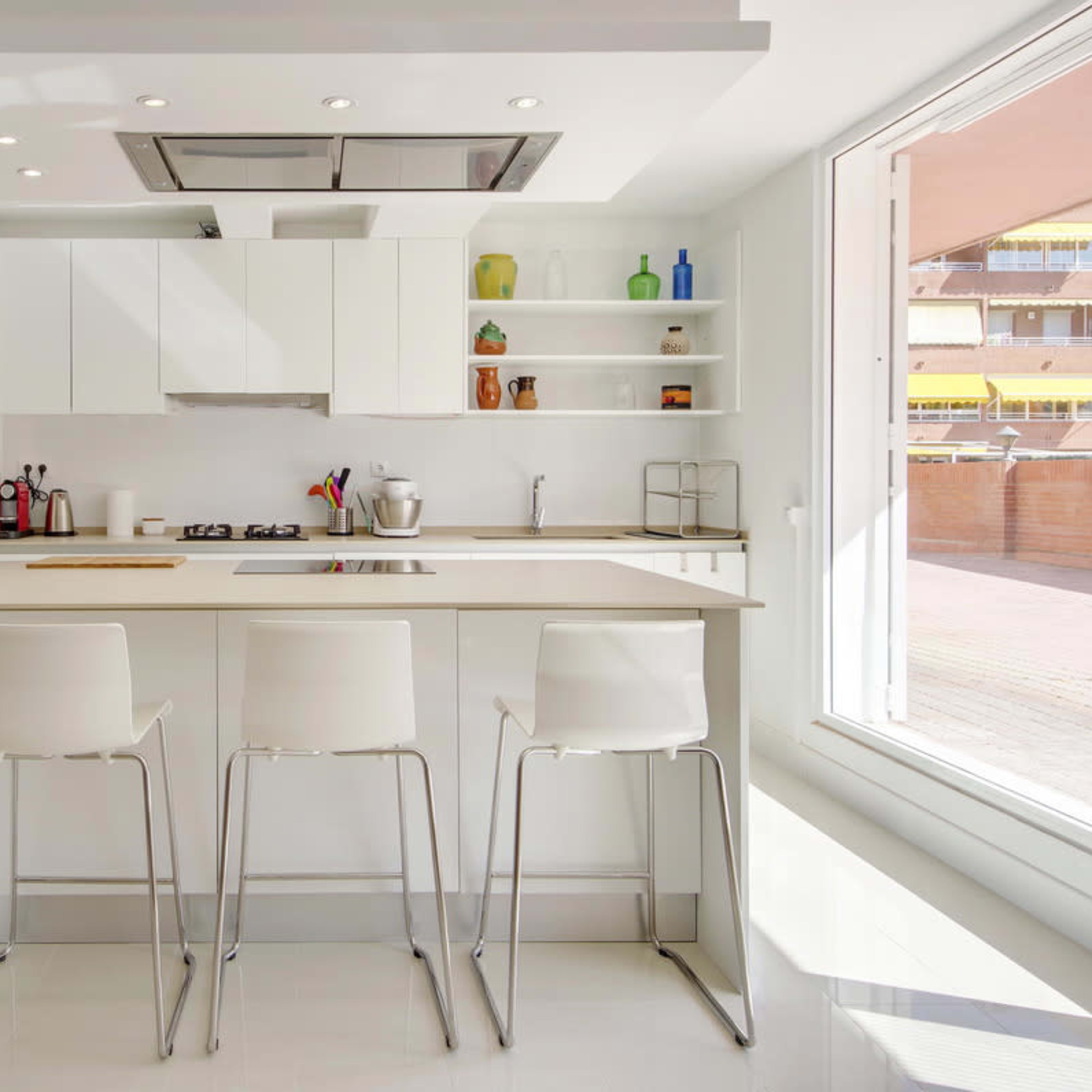 A modern kitchen features a central island with four white bar stools, light countertops, and sleek cabinetry against a bright, airy background.