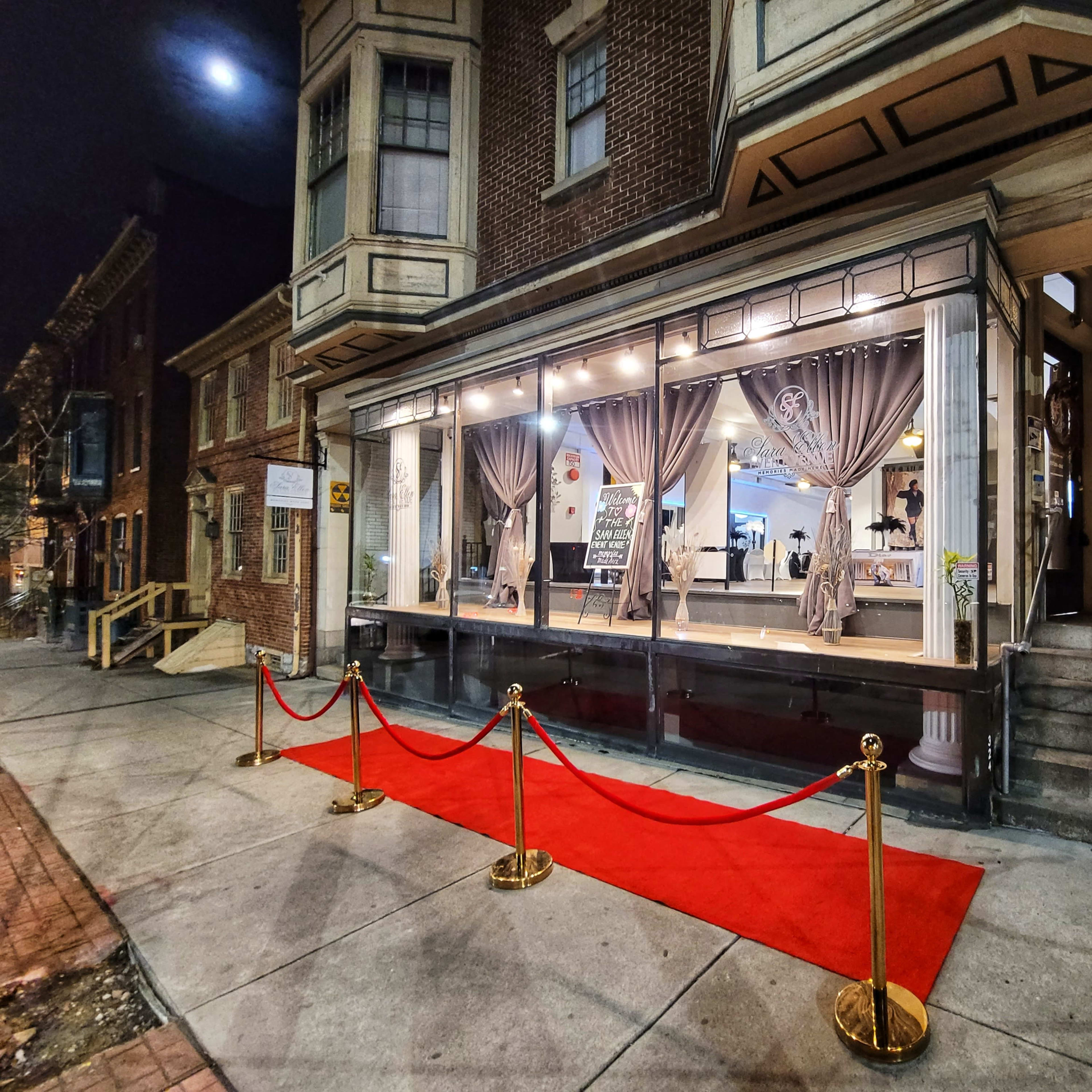 A red carpet lined with stanchions leads to a storefront with large glass windows, showcasing an interior decorated with curtains and seating arrangements, set against a backdrop of brick buildings at night.