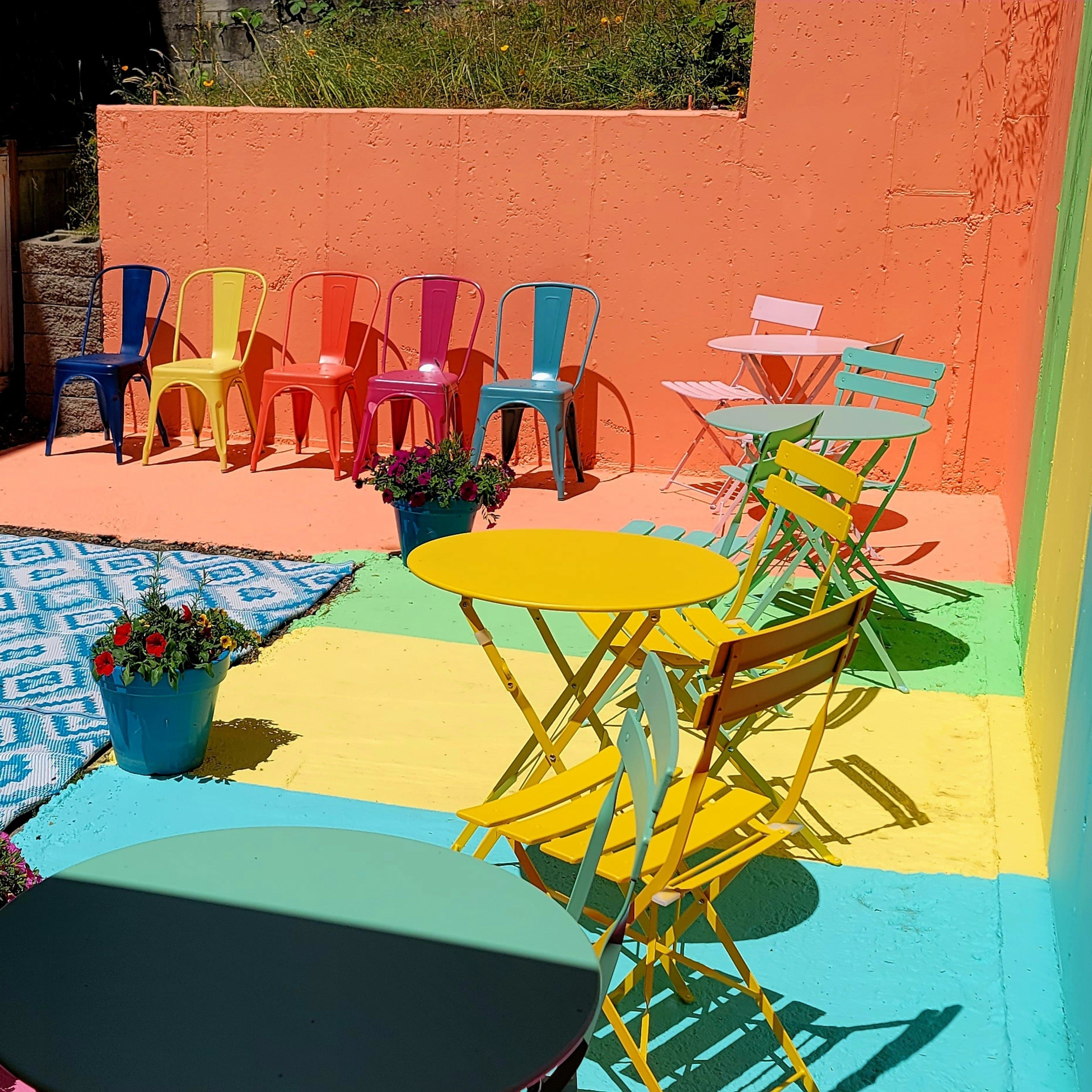 The scene features a vibrant outdoor area with colorful seating, including yellow and teal tables and multicolored chairs, surrounded by potted flowers and a patterned blue rug.
