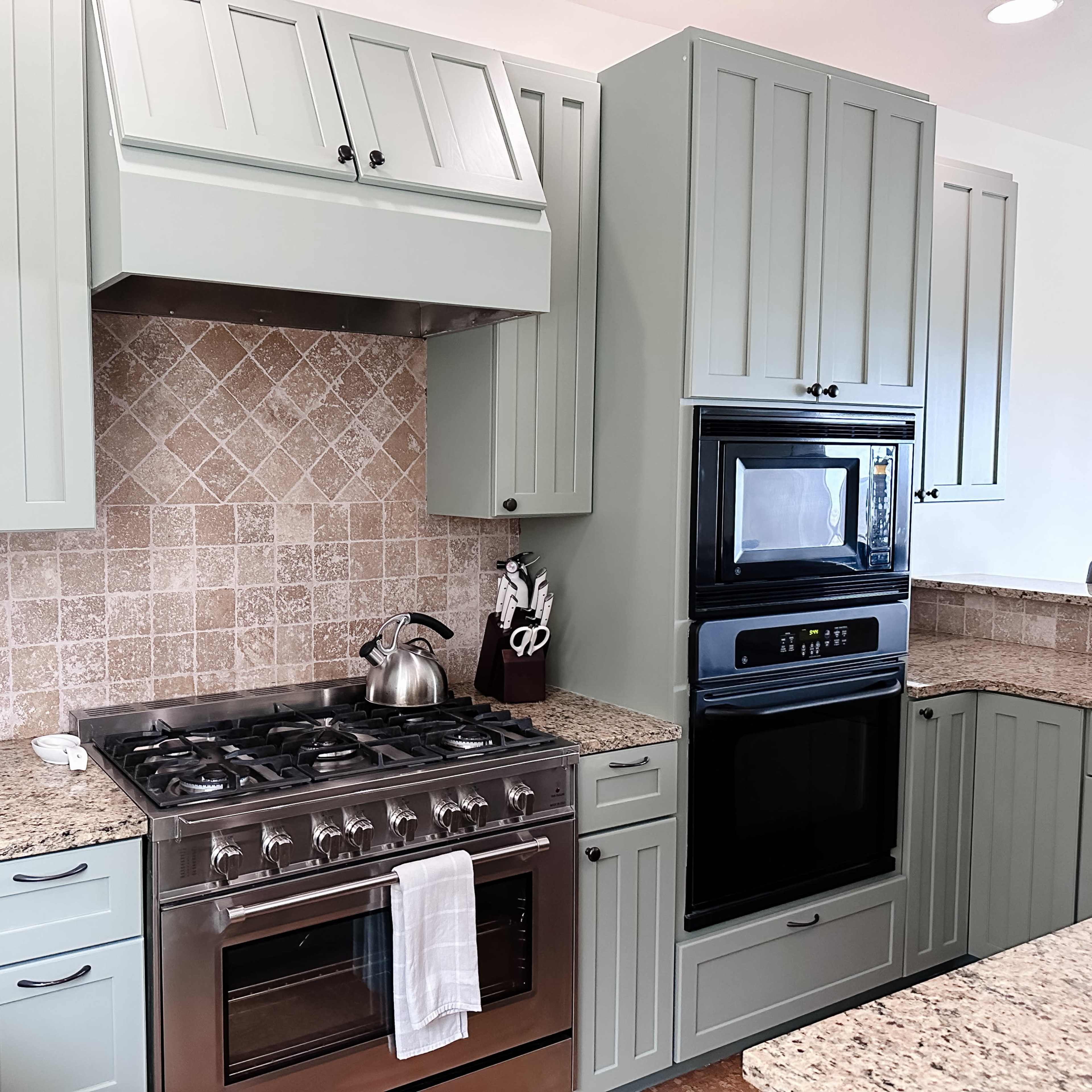 A modern kitchen features green cabinetry, a stainless steel gas stove, a black built-in oven, and granite countertops with a tiled backsplash.