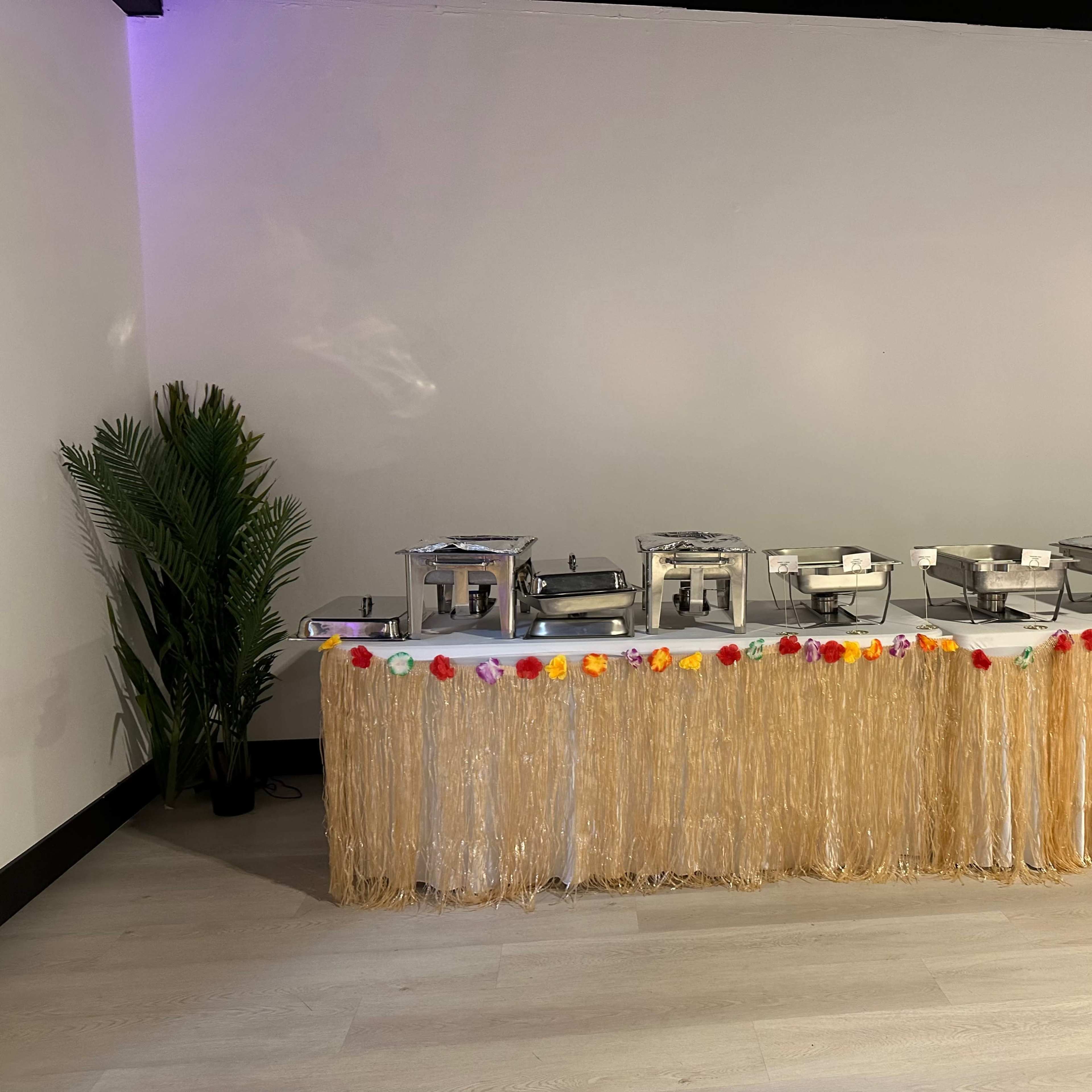 A table decorated with a fringed skirt displays several chafing dishes, flanked by potted palm plants.