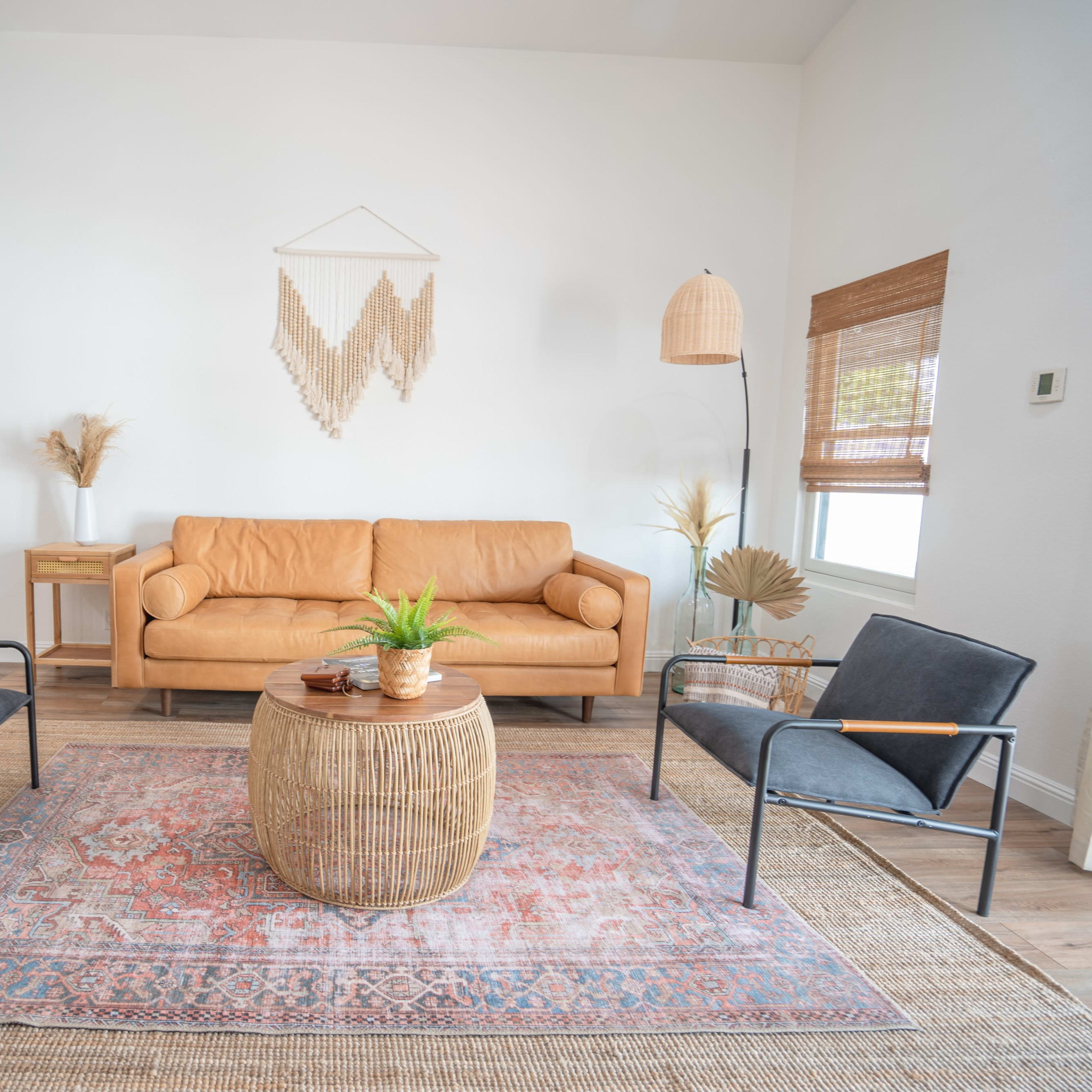 A stylish living room features a leather couch, two black chairs, a round woven coffee table, and a decorative rug on a wooden floor.