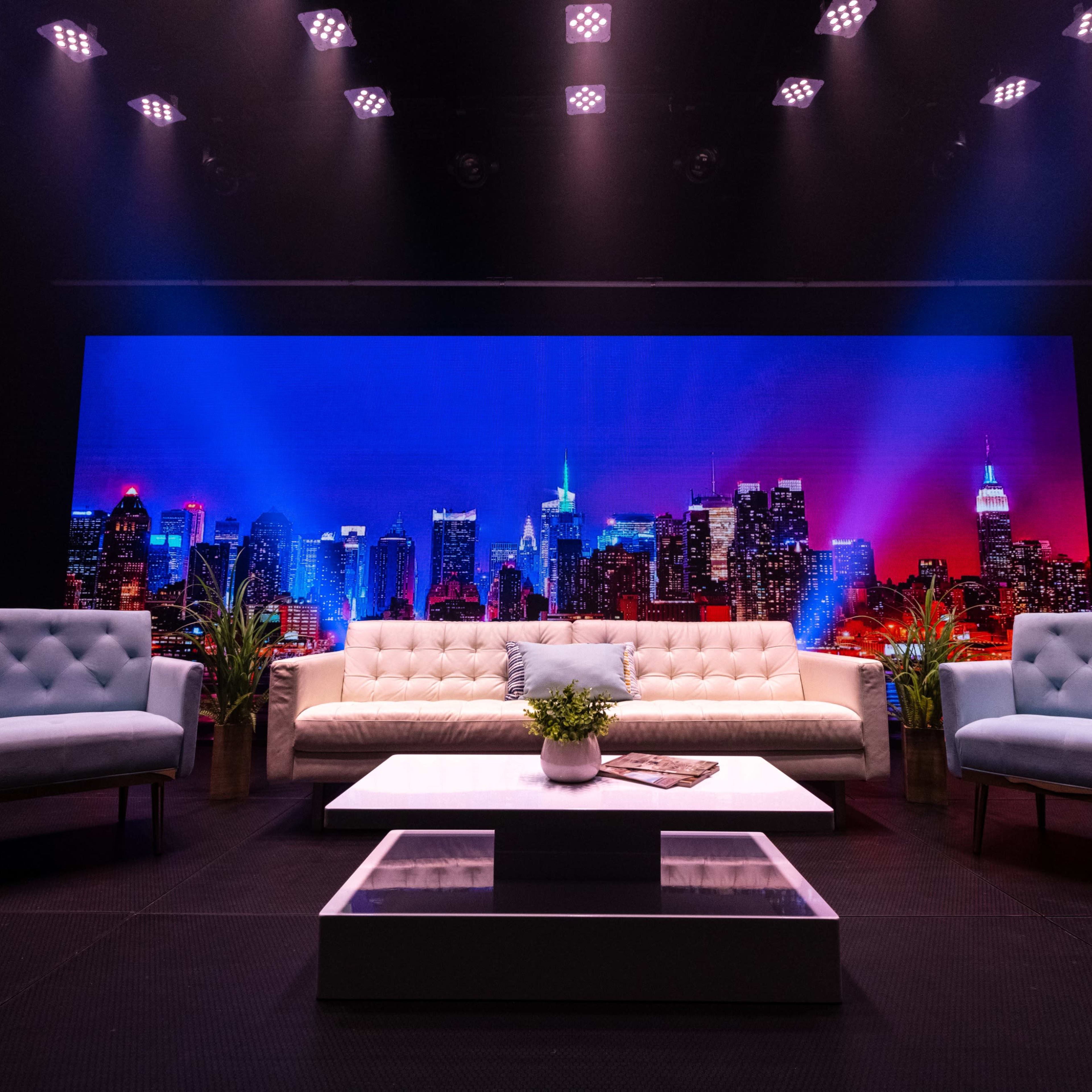 A modern stage setup with a white sofa and two light blue armchairs facing a large backdrop of a city skyline illuminated in blue and purple hues.