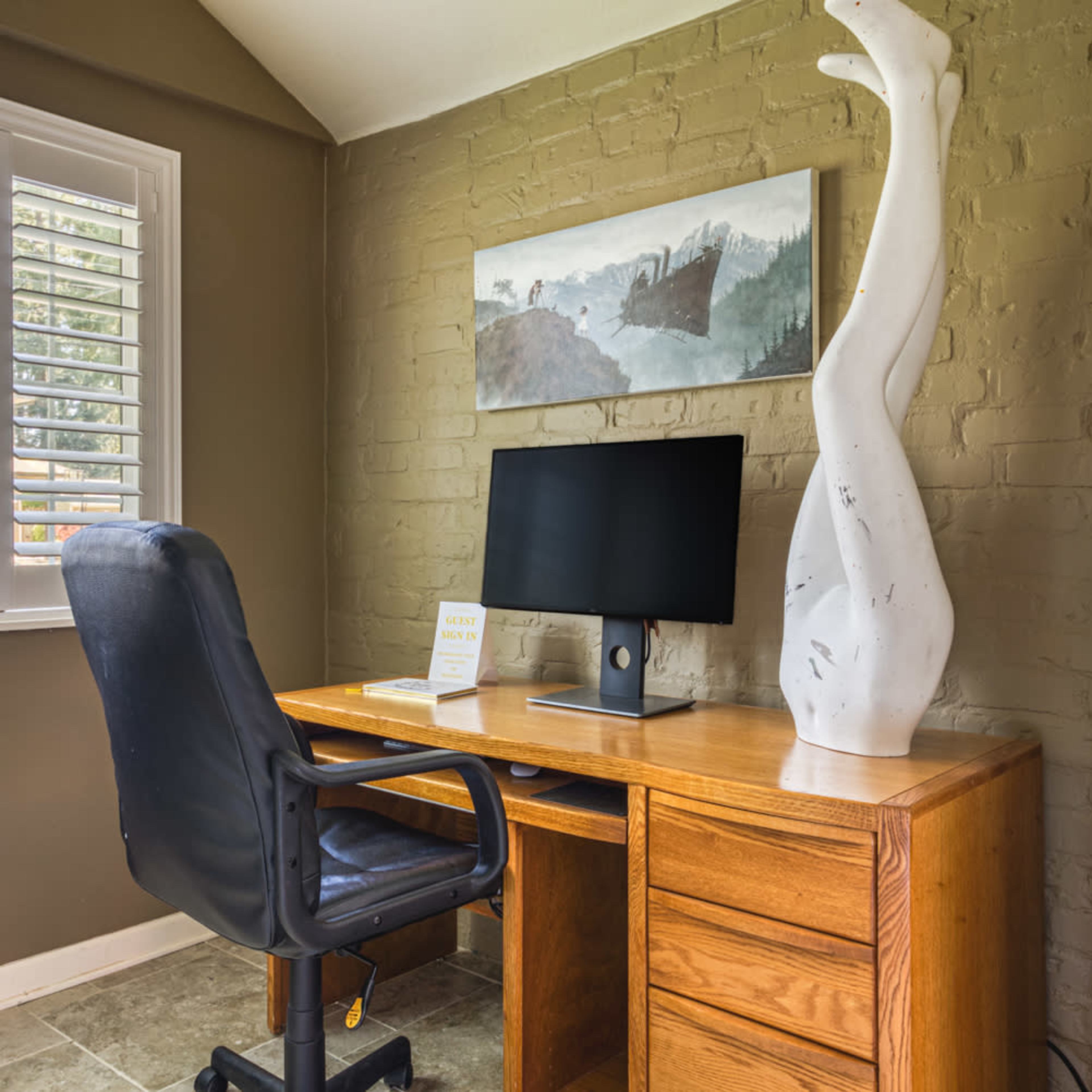 A home office features a wooden desk, an office chair, a computer monitor, and a decorative sculpture on the surface.