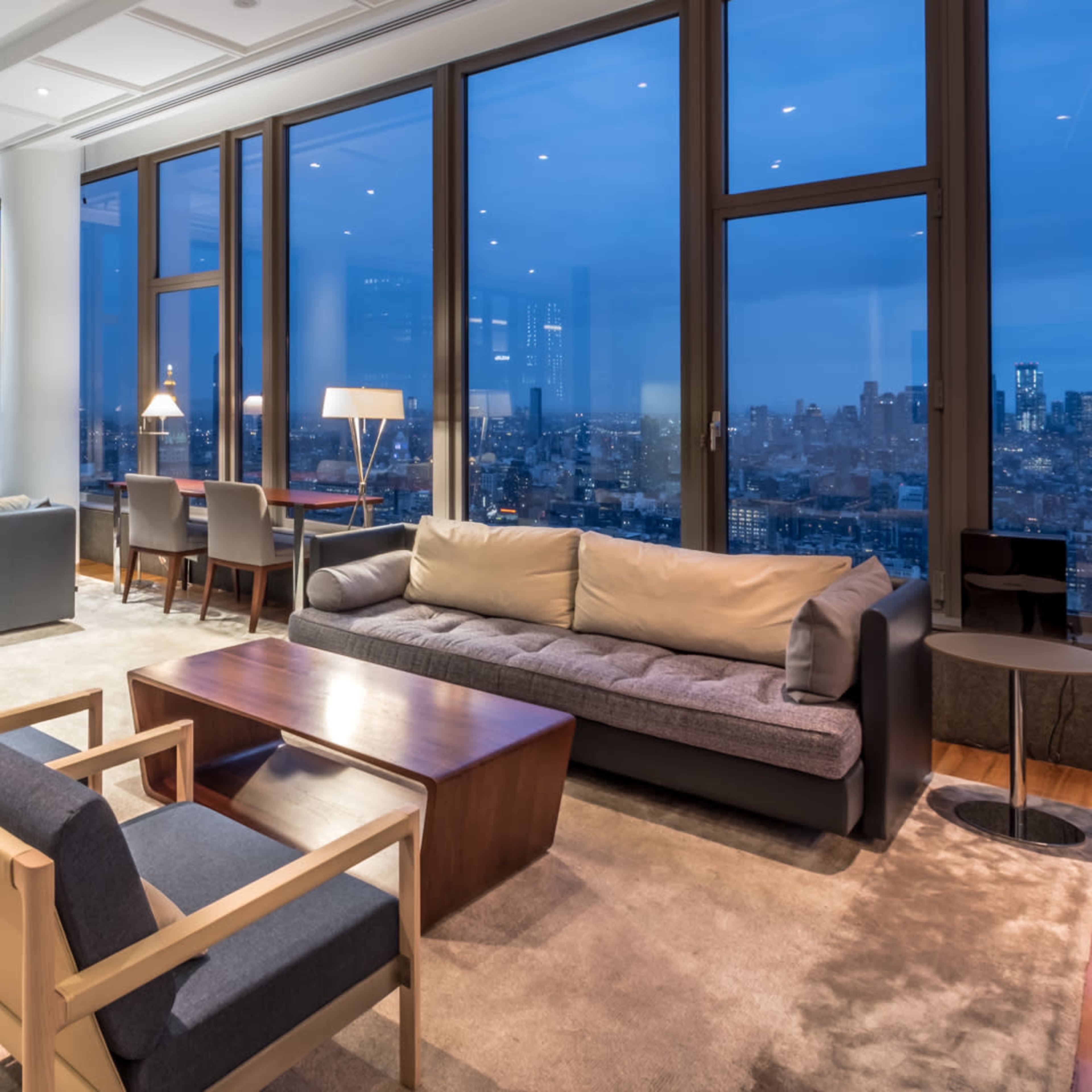 A modern living room with large windows offering a view of a city skyline at dusk, featuring minimalist furniture and soft lighting.