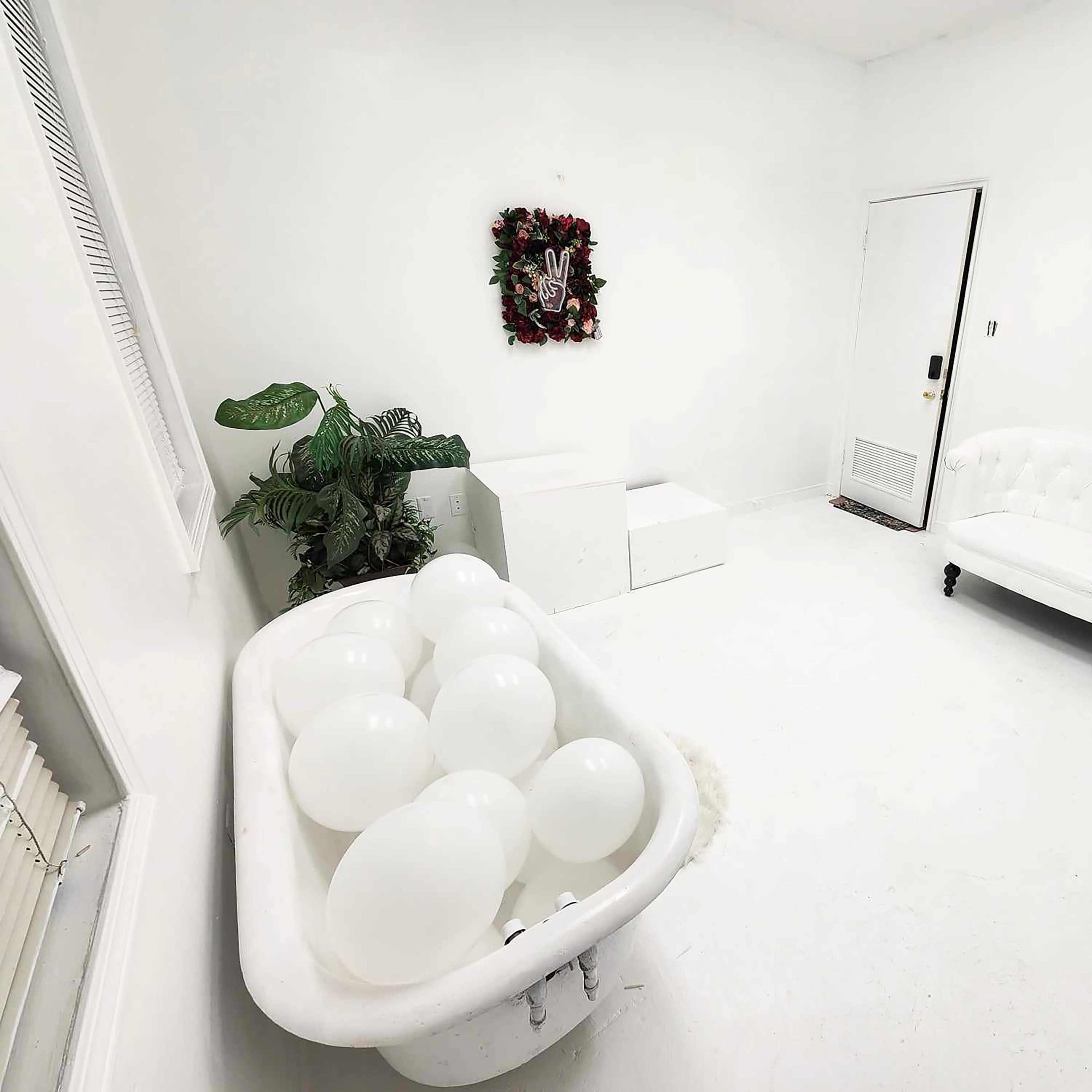 A bright white room features a vintage bathtub filled with white balloons, a white couch, a small table, and potted plants.