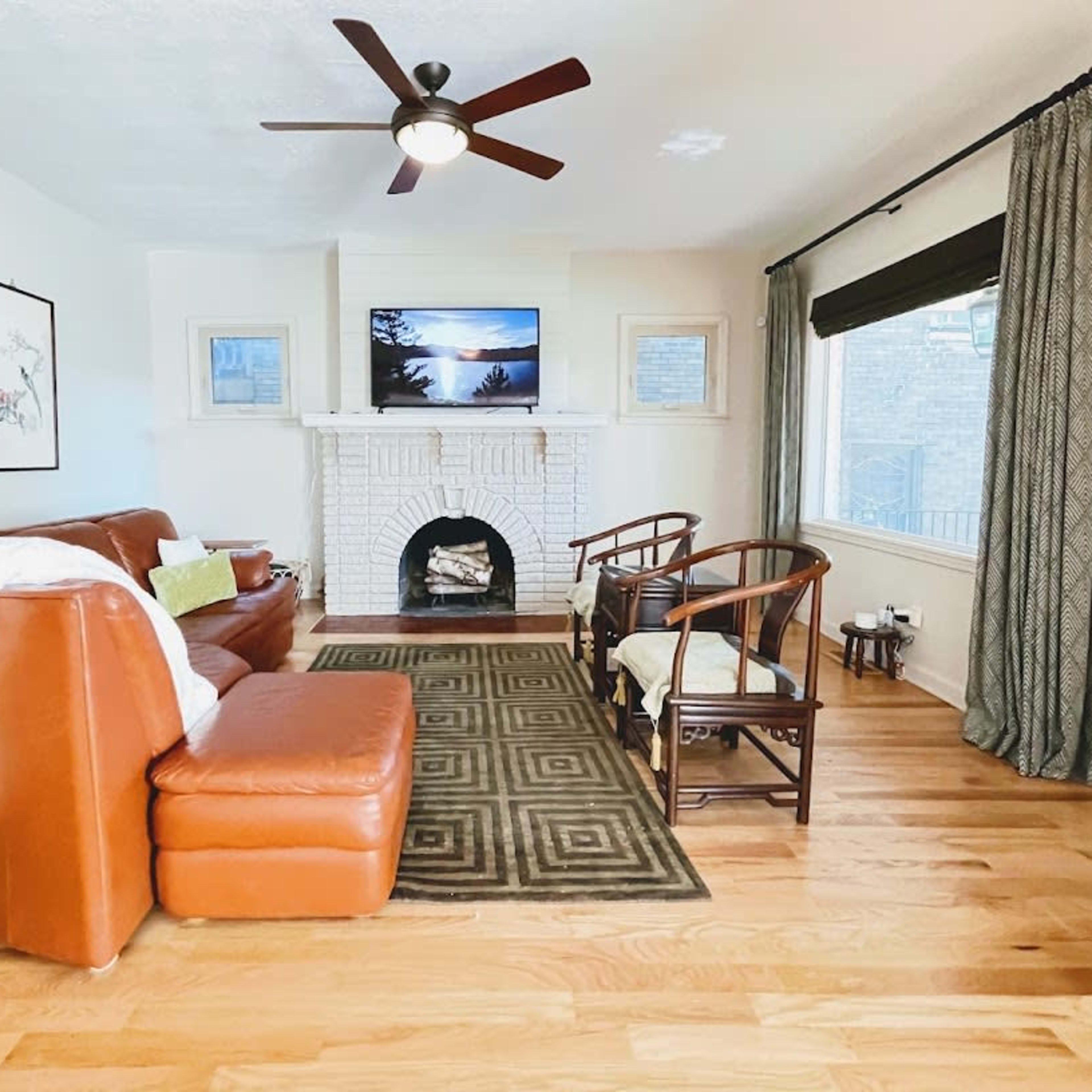 A living room features a brown leather sofa, two wooden chairs, a white brick fireplace, and a television mounted on the wall.