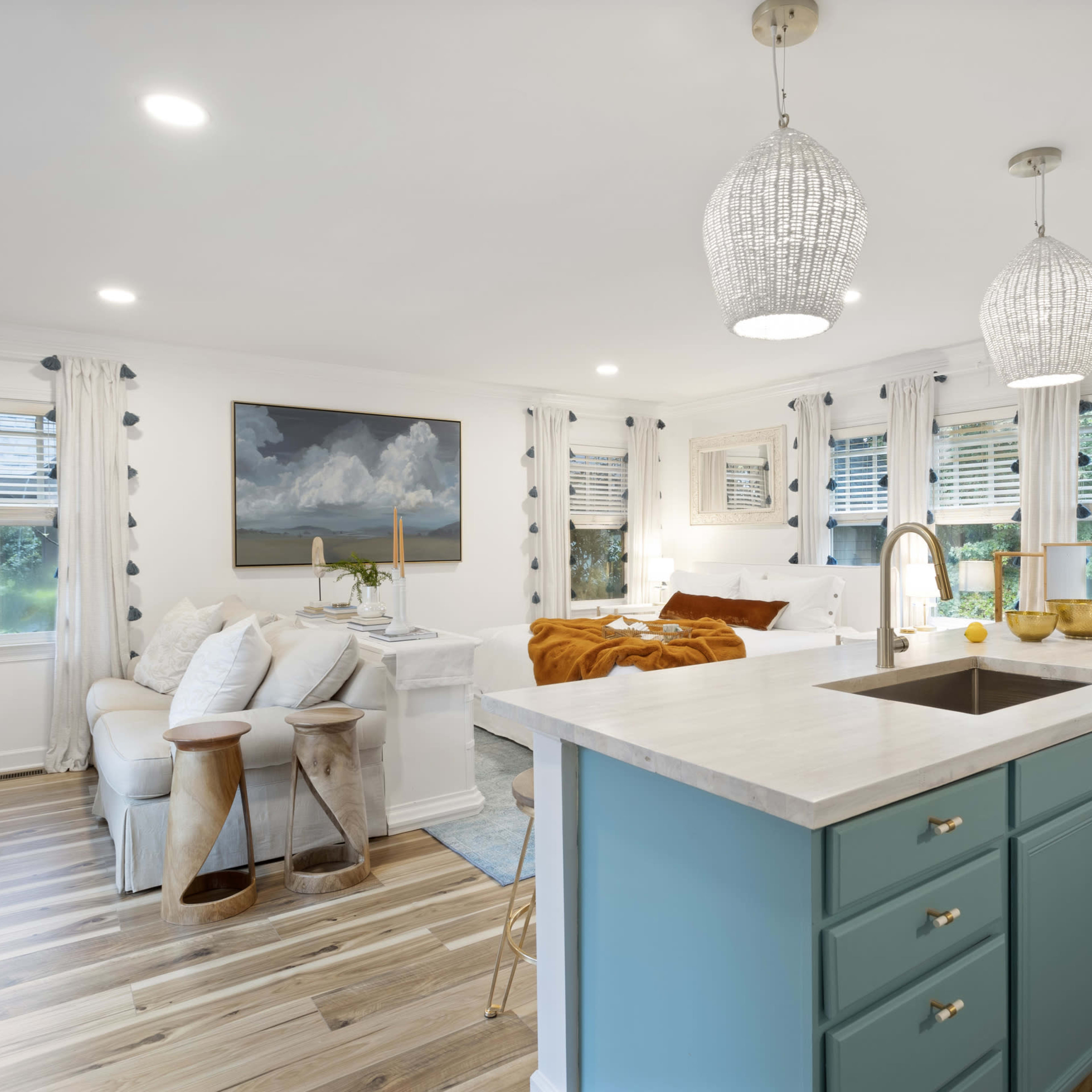 A modern kitchen and living area featuring a central island with a marble countertop, teal cabinetry, and bright natural light streaming through multiple windows.