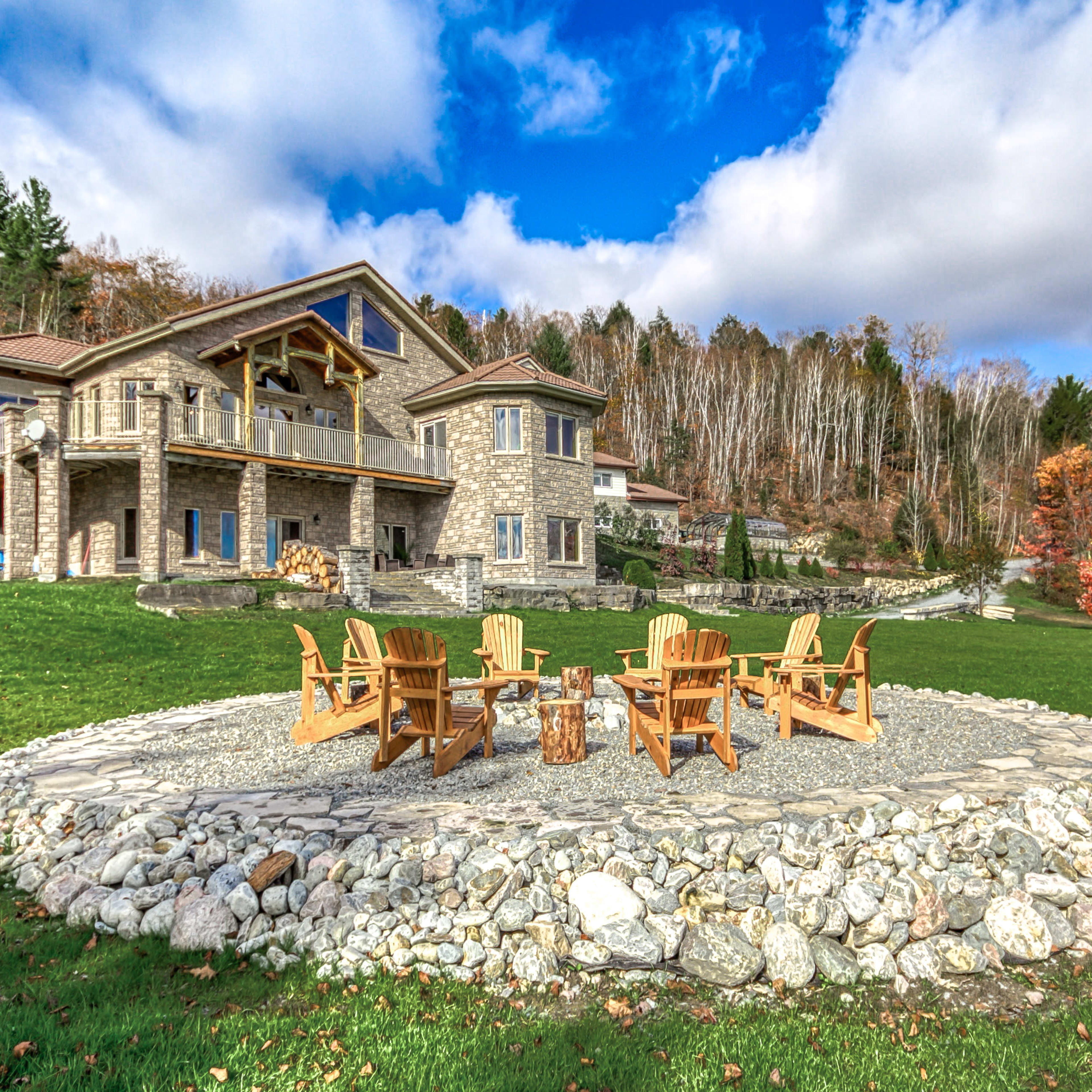 A stone patio with a circular seating arrangement of wooden chairs is situated in front of a large stone house, surrounded by a lush green lawn and trees.