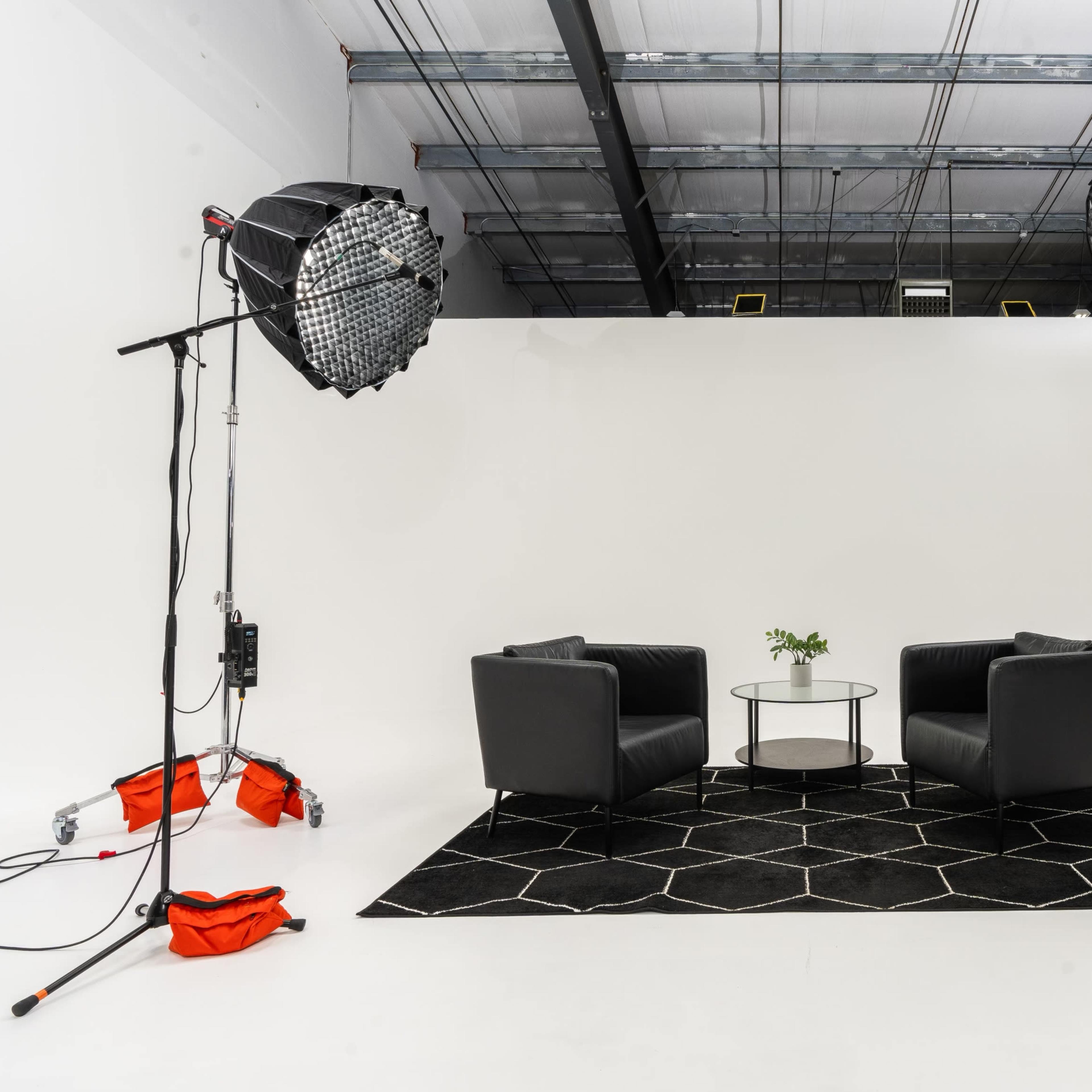 A minimalist photography studio setup with two black chairs facing each other, a glass table in the center, and large softbox lights positioned on either side.