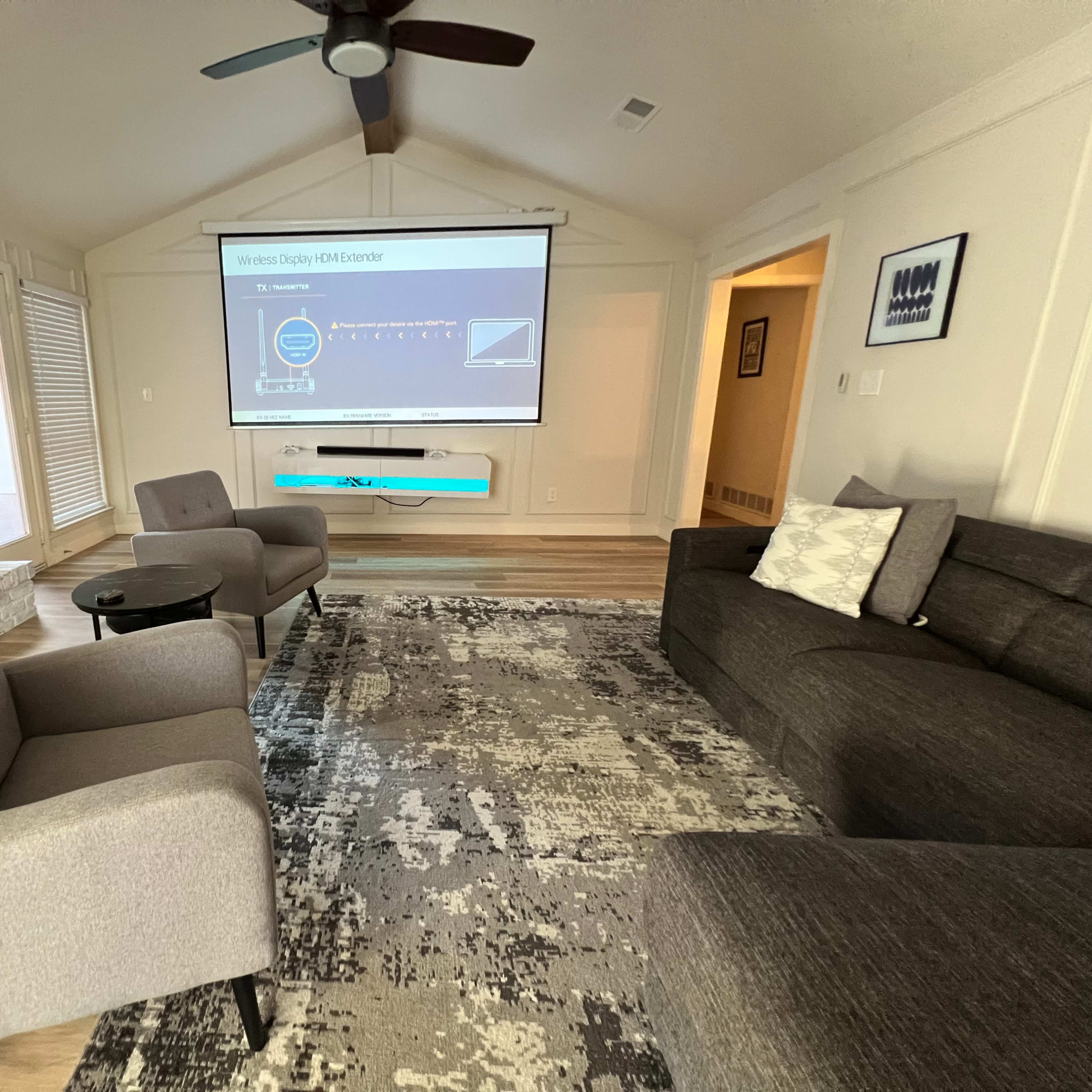 A modern living room with a large screen displaying a presentation, two gray upholstered armchairs, a gray sofa, and a patterned area rug on wooden flooring.