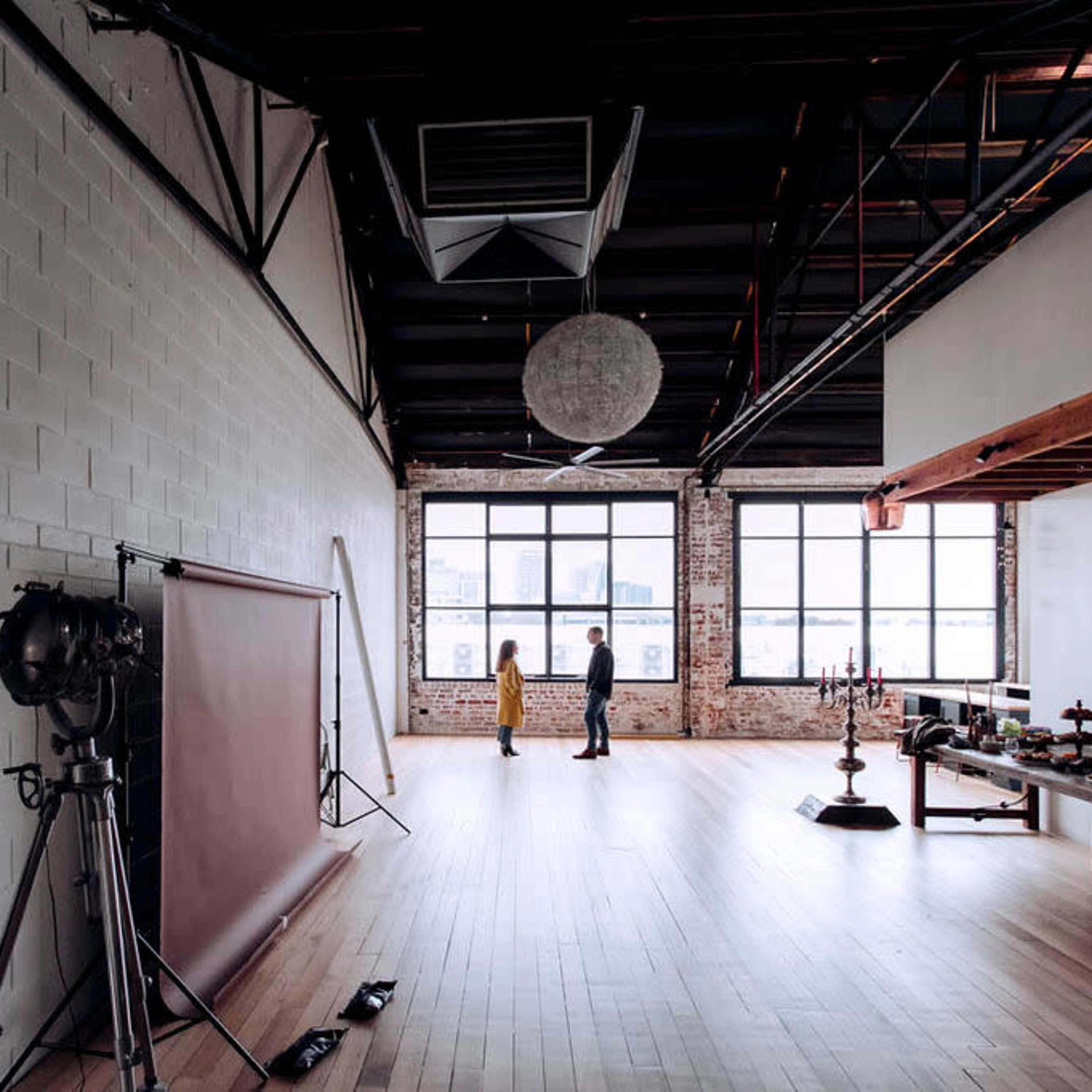 Two people stand in a spacious, industrial-style room with large windows and wooden flooring, surrounded by photography equipment and furnishings.