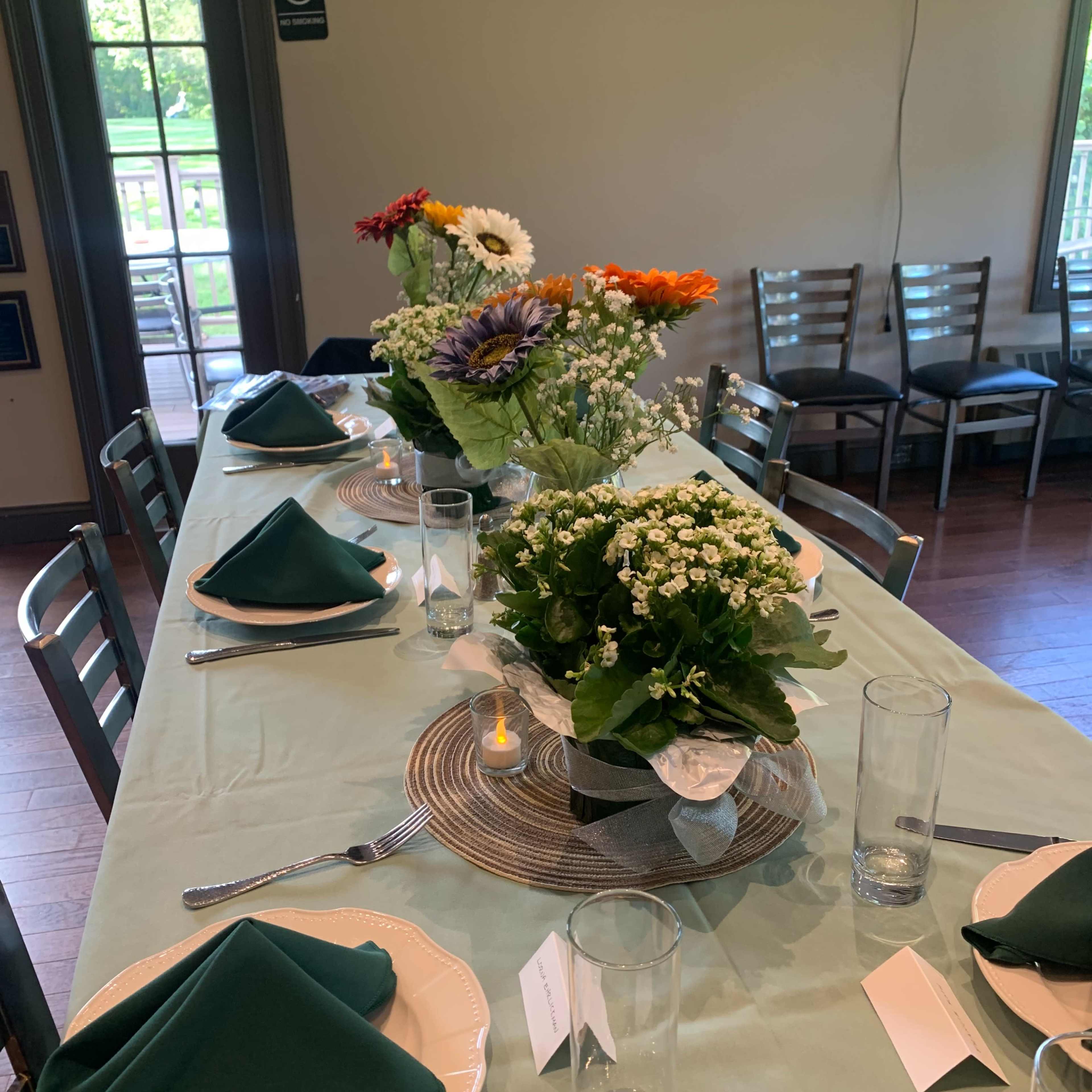 A neatly arranged long dining table is set with green napkins, clear glasses, and centerpieces of flowers and plants, in a well-lit room with wooden flooring.