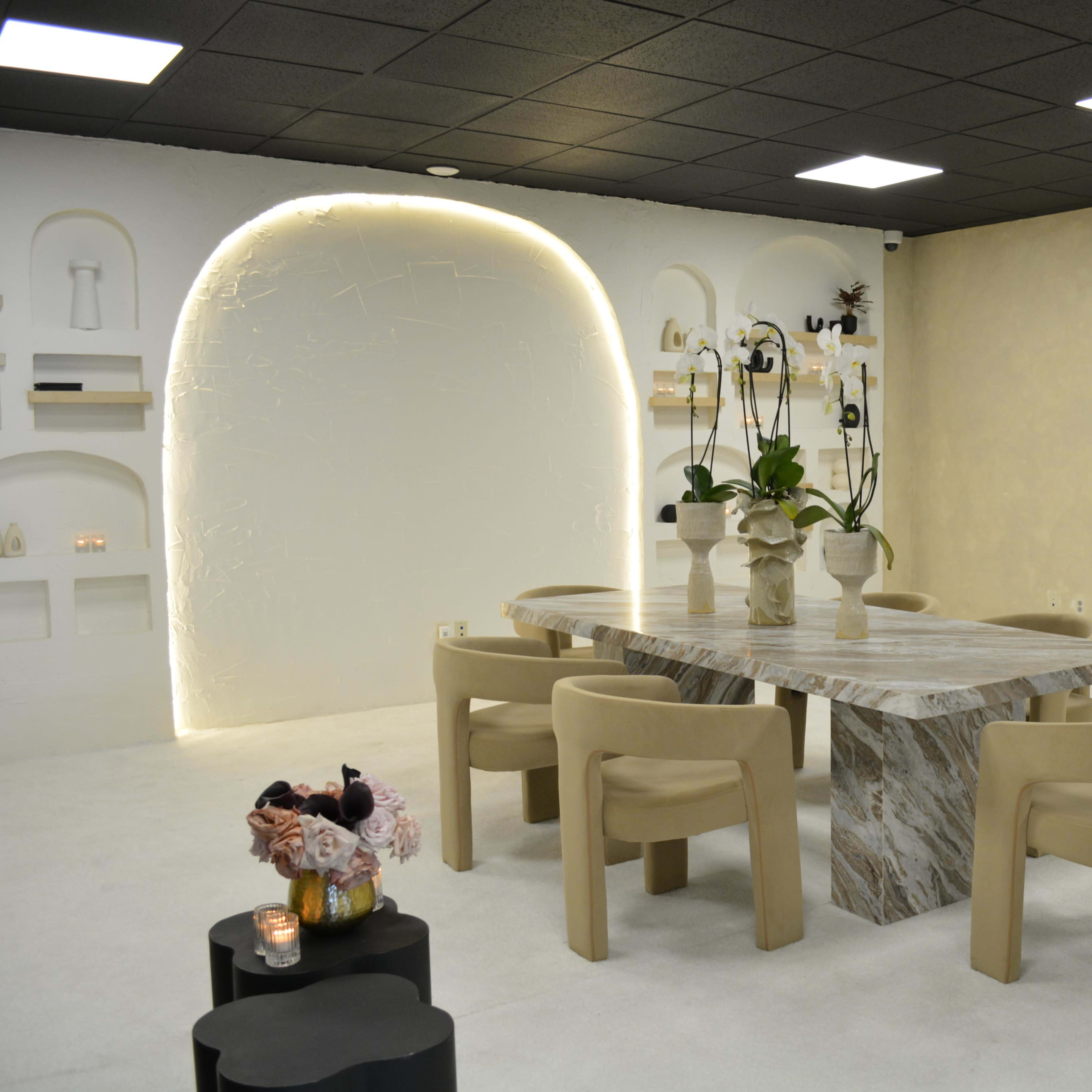 A modern dining room with a marble table, beige chairs, and decorative shelving against softly lit walls.