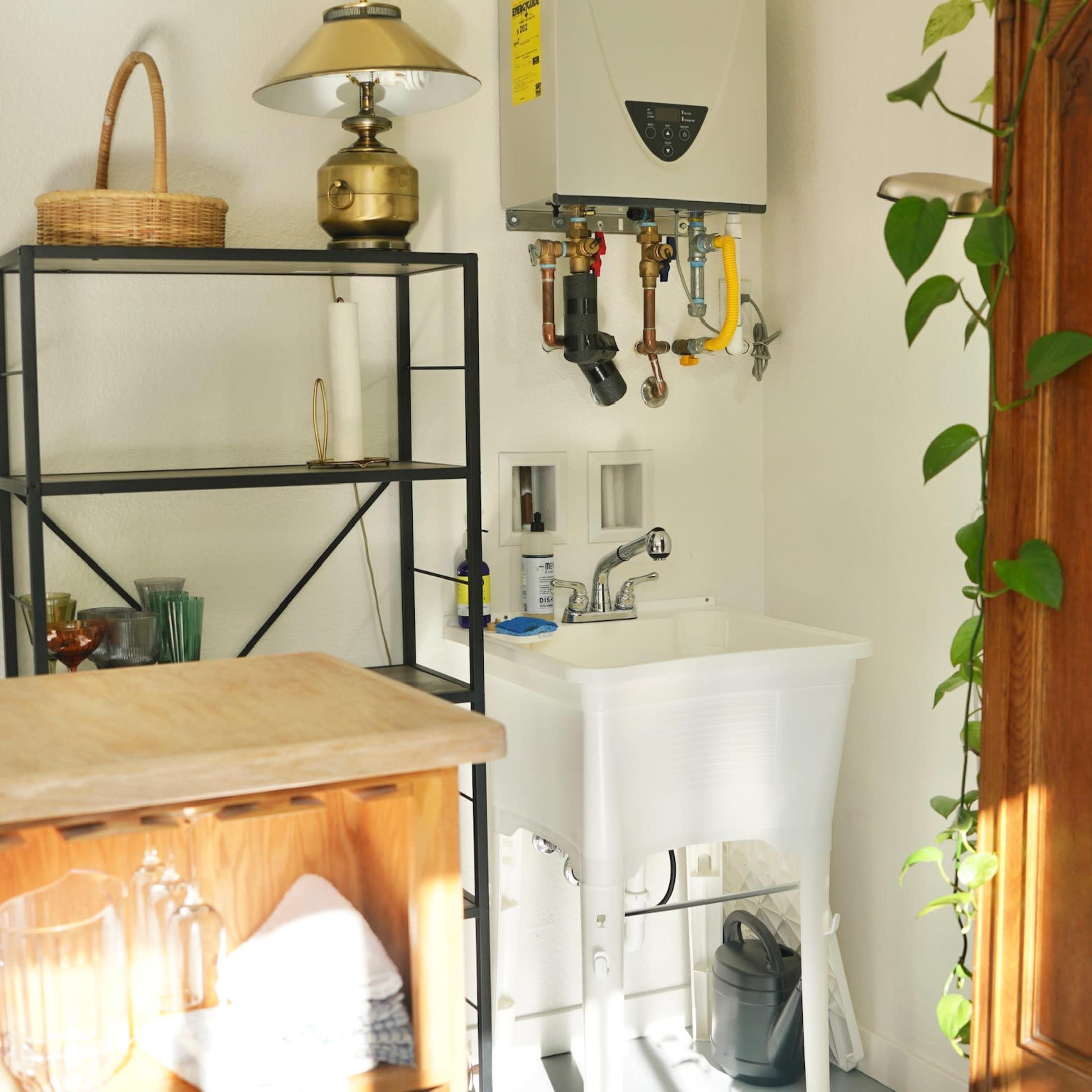 A small utility area featuring a wall-mounted water heater, a sink, and a shelving unit with a lamp and various items.