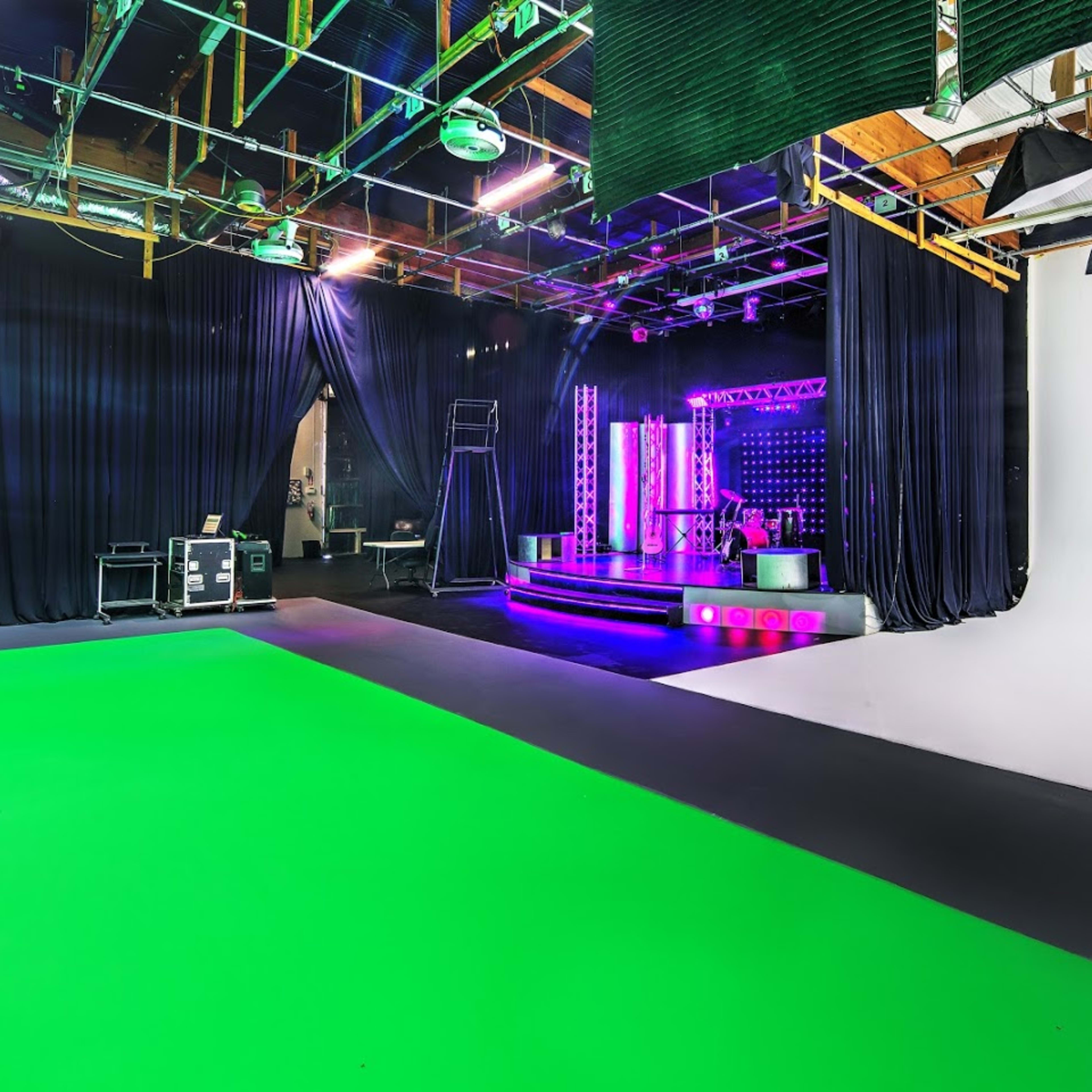 A production studio with a green screen area, black drapes, and a stage featuring colorful lights and equipment in the background.
