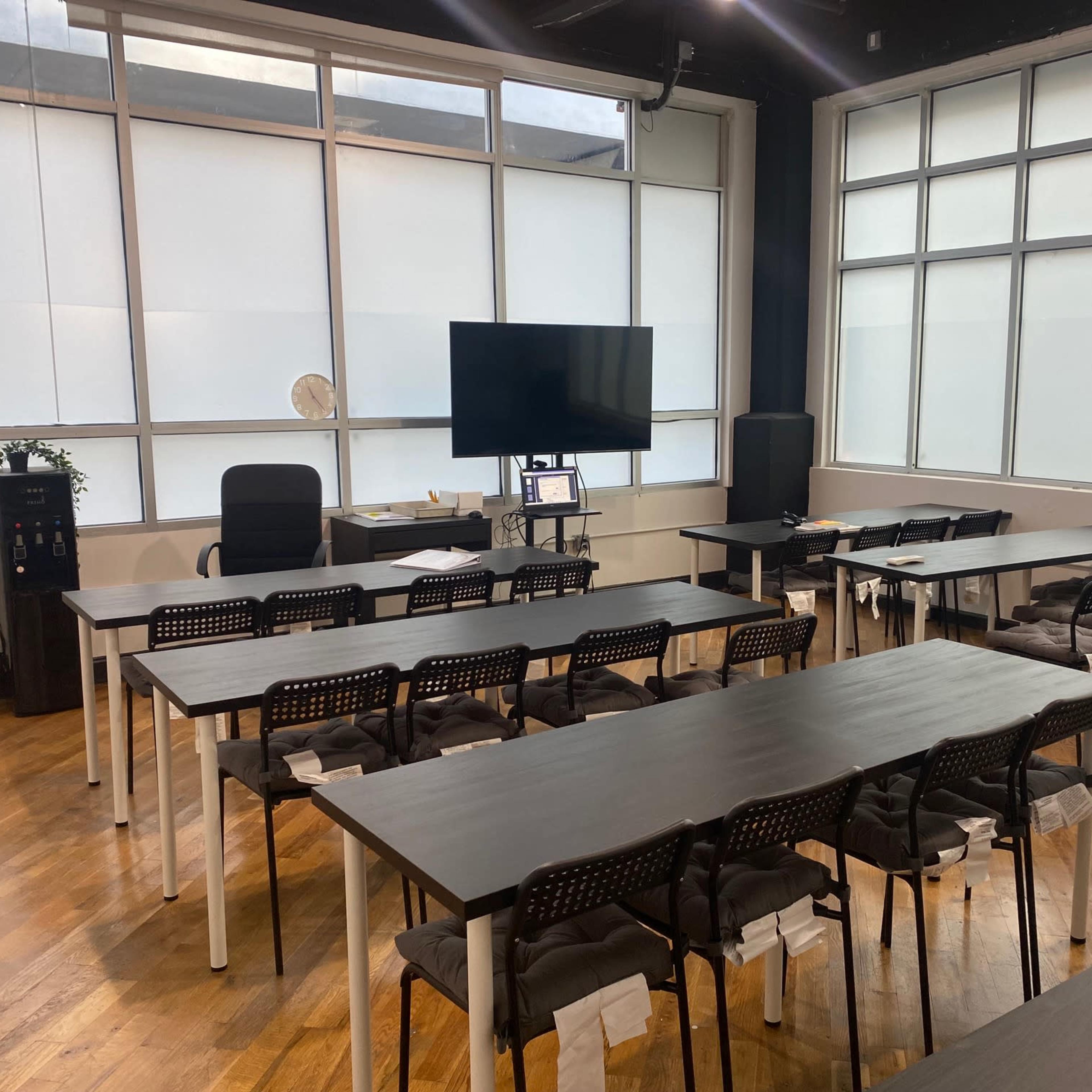 A well-lit classroom or meeting space with several tables and chairs arranged in a dual-row layout, a large screen on the wall, and a desk in the background.