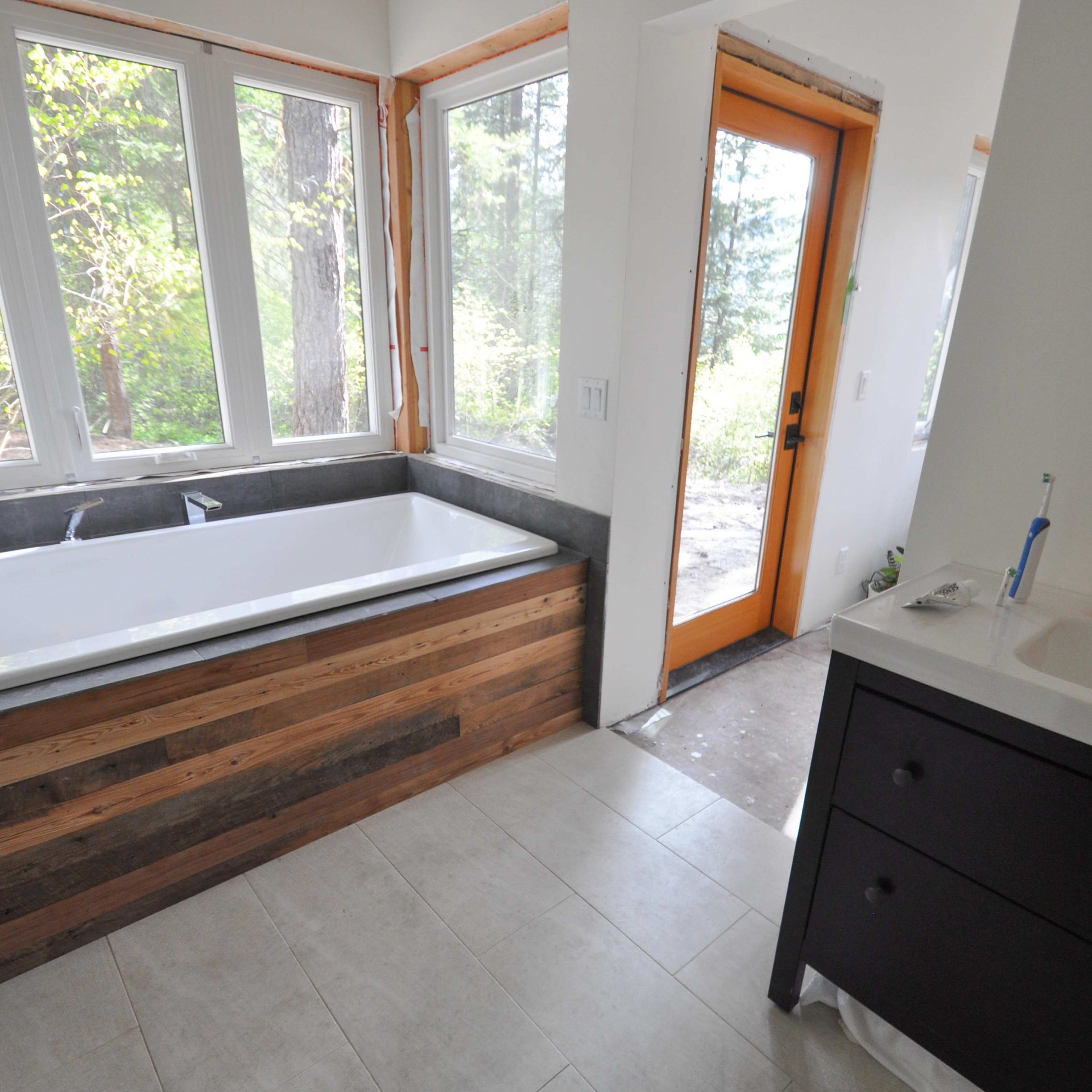 A modern bathroom with a wooden bathtub surround, a sink cabinet, and a door leading outside to a natural setting.
